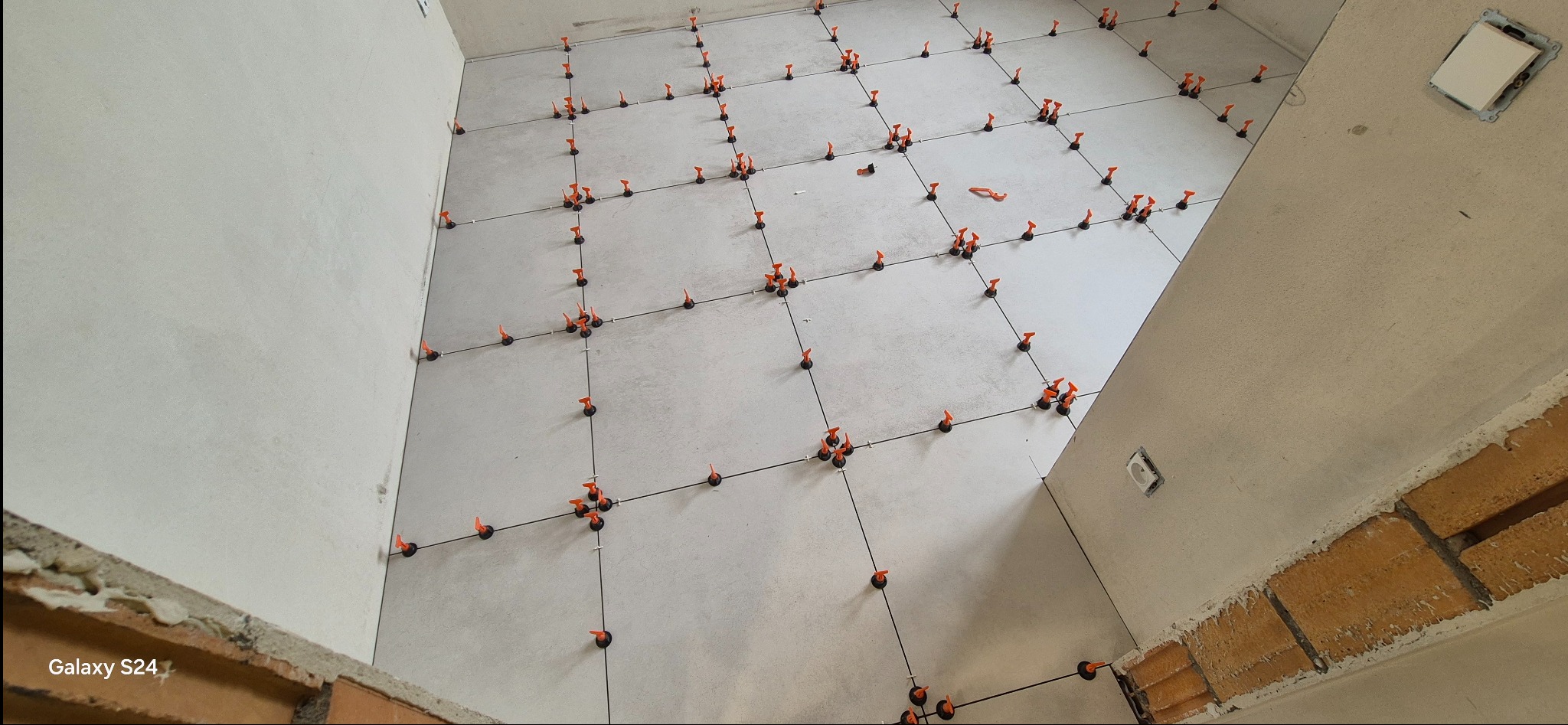 Jasne płytki podłogowe w trakcie układania, z pomarańczowymi klipsami poziomującymi i czarnymi krzyżykami dystansowymi. Widok z góry, surowy stan wykończenia ścian.