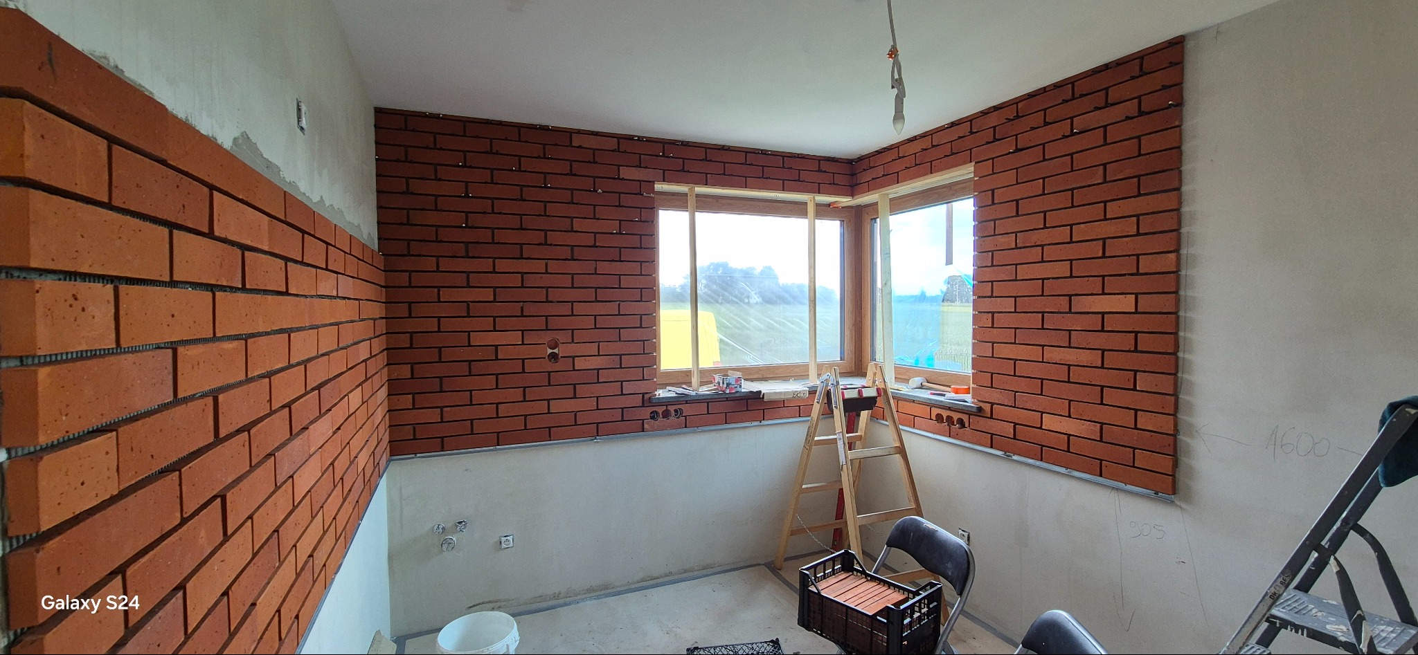 Wnętrze w trakcie prac: częściowo wykończone ściany z cegły klinkierowej, surowy tynk na dole, okno z widokiem na zewnątrz, drabina i narzędzia w pomieszczeniu.