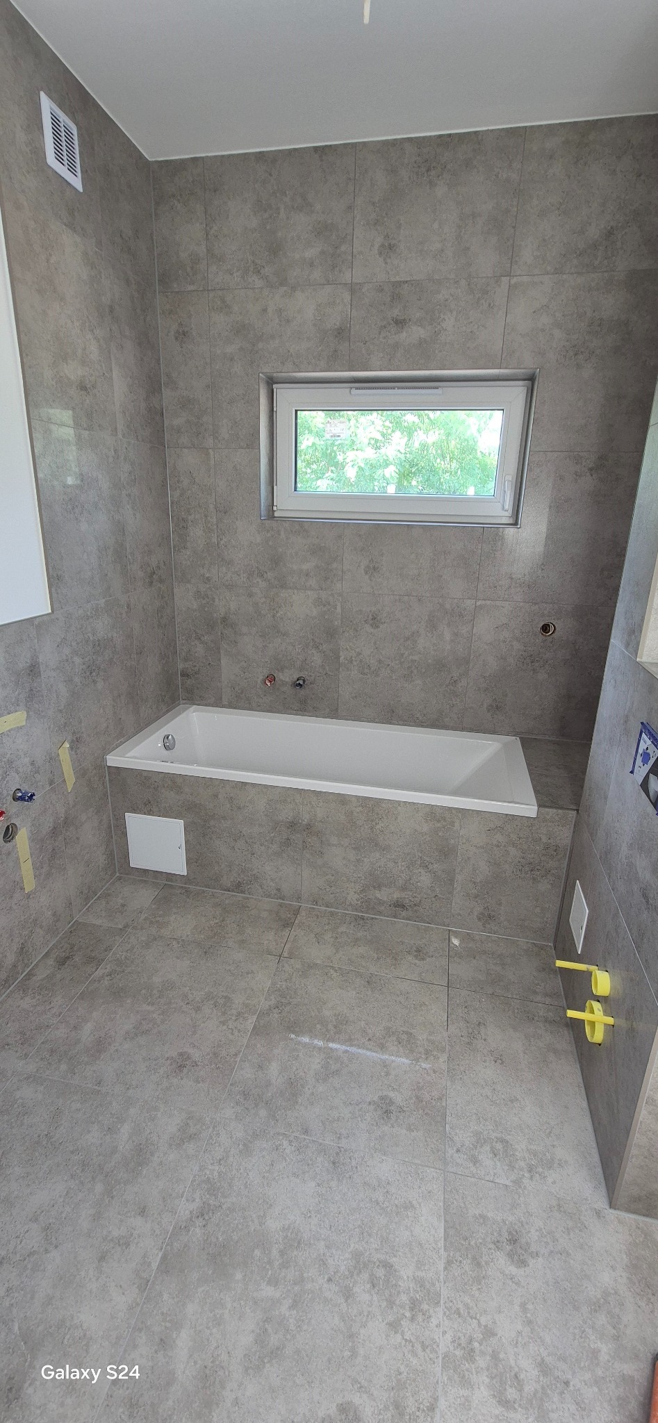 Wykończona łazienka w szarościach z wanną pod oknem. Widoczne płytki na ścianach i podłodze oraz elementy instalacji wodnej. Minimalistyczny design.