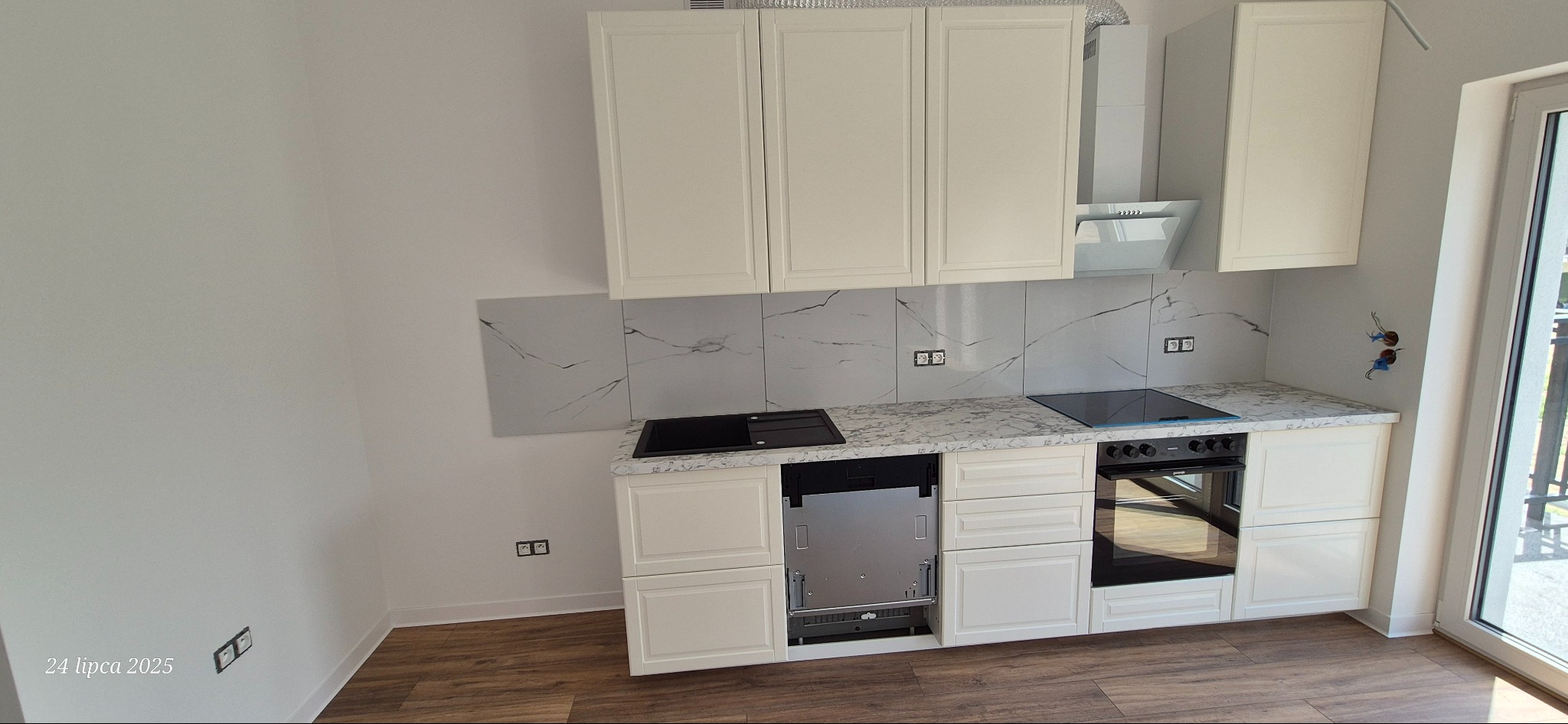 Jasna kuchnia z białymi szafkami i marmurowymi płytkami. Widoczna płyta indukcyjna, piekarnik i okap. Nowoczesne wykończenie wnętrza z drewnianą podłogą.