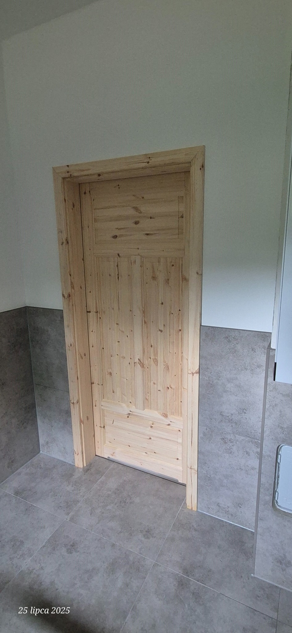 Surowe drewniane drzwi z ościeżnicą w nowej łazience z szarymi płytkami podłogowymi i ściennymi, widok na fragment ściany i podłogi, naturalne światło dzienne.