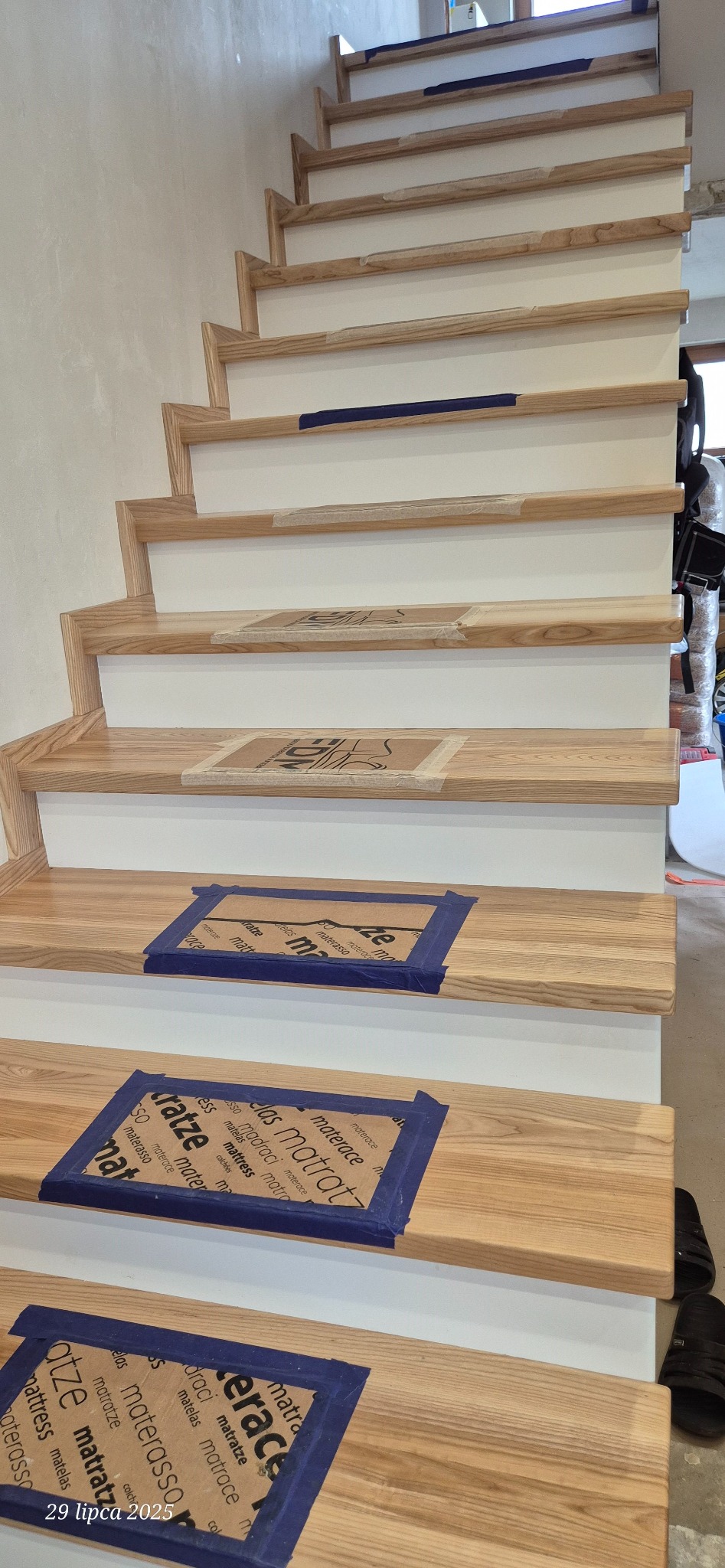 Drewniane schody z białymi podstopnicami w trakcie montażu, zabezpieczone kartonem i taśmą malarską. Widoczne fragmenty ściany i podłogi.