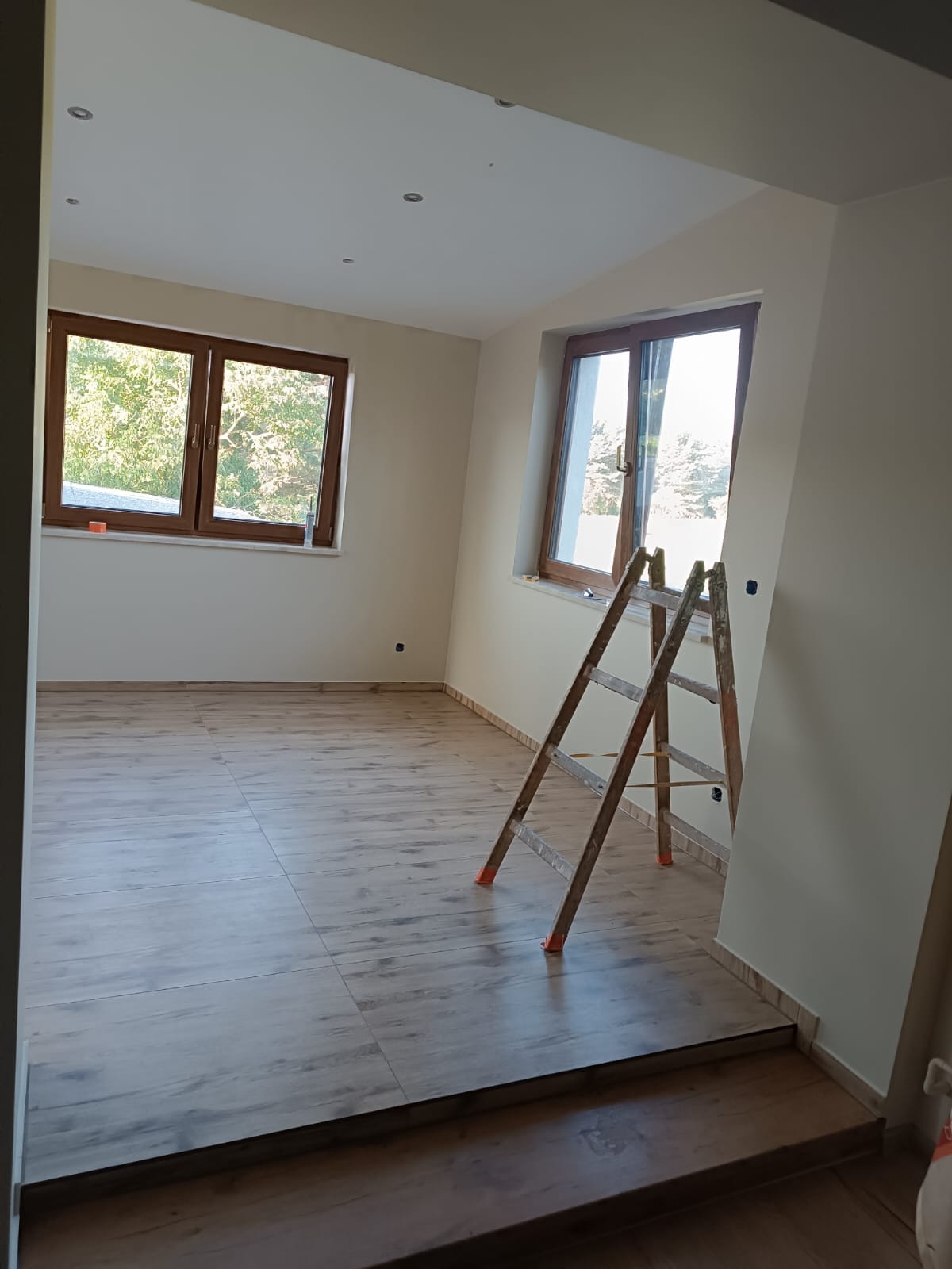 Jasne wnętrze po remoncie z drewnopodobną podłogą, drabina oparta o świeżo pomalowaną ścianę, okna z widokiem na zieleń. Widoczne nowe oświetlenie sufitowe.