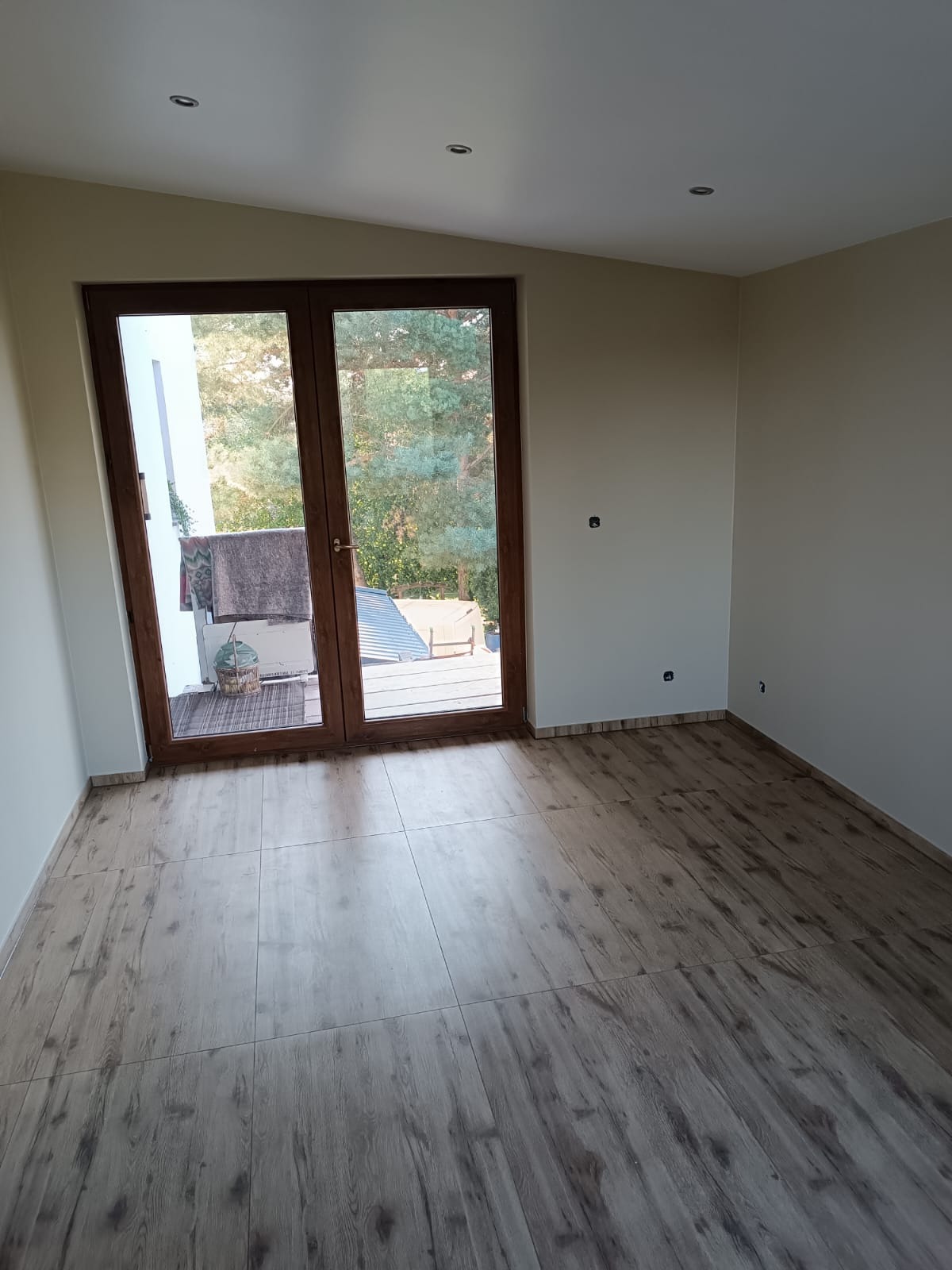 Jasne wnętrze po remoncie z panelami podłogowymi w odcieniach drewna i dużym oknem balkonowym z widokiem na zieleń. Widoczne listwy przypodłogowe.