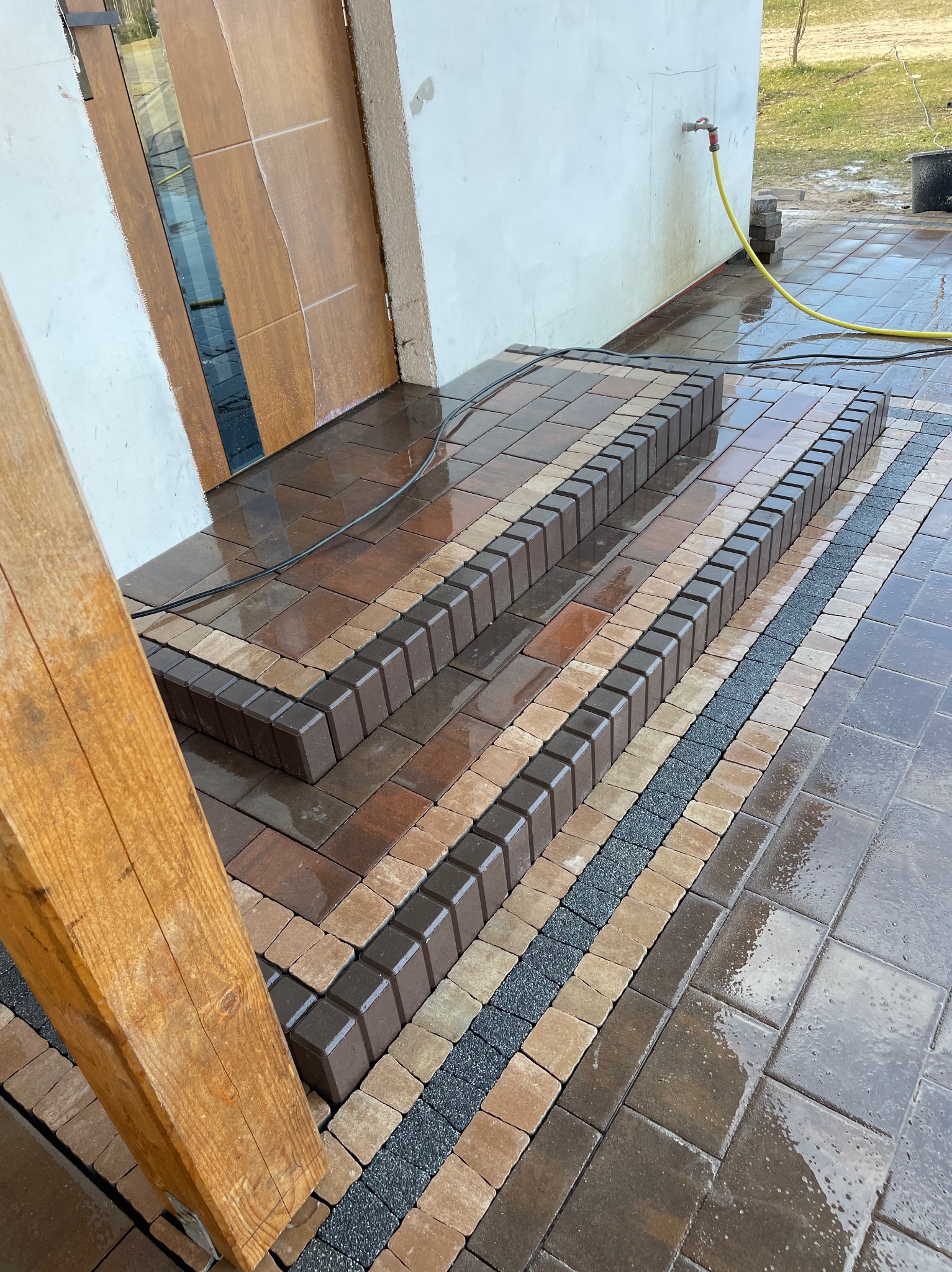 Ułożone schody z kostki brukowej w odcieniach brązu i beżu, oddzielone pasem drobnego grysu, prowadzące do drzwi wejściowych. Powierzchnia mokra, prawdopodobnie po myciu.