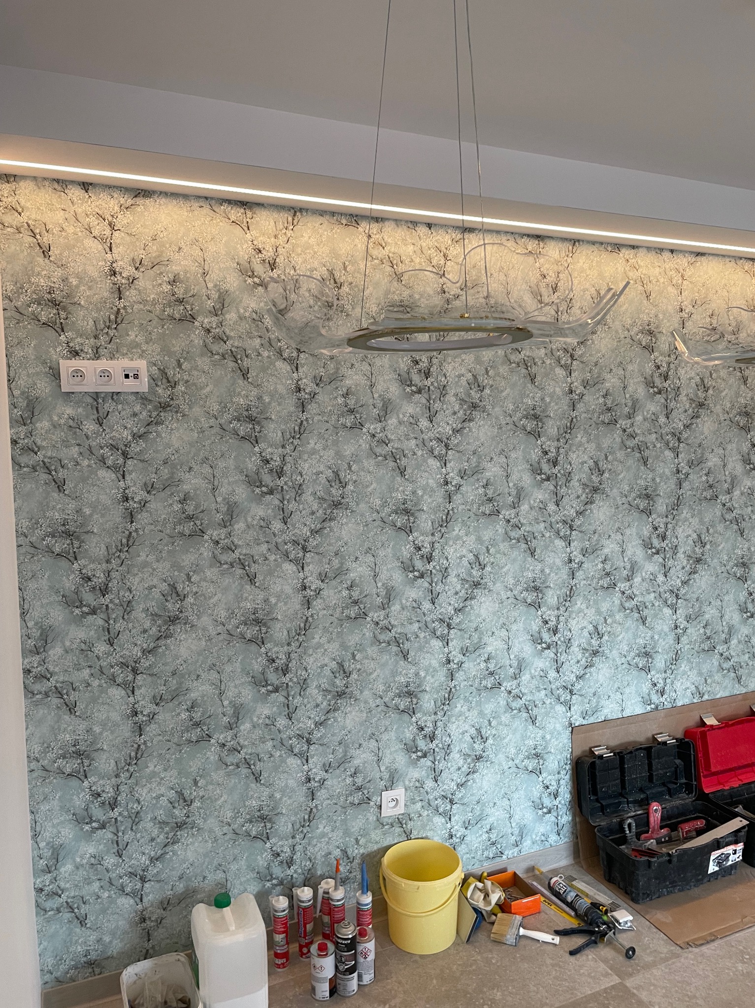 Ściana pokryta tapetą w motywy drzew, z widocznymi gniazdkami elektrycznymi, lampą wiszącą i narzędziami malarskimi oraz pojemnikami z masami uszczelniającymi na podłodze, sugerującymi prace...