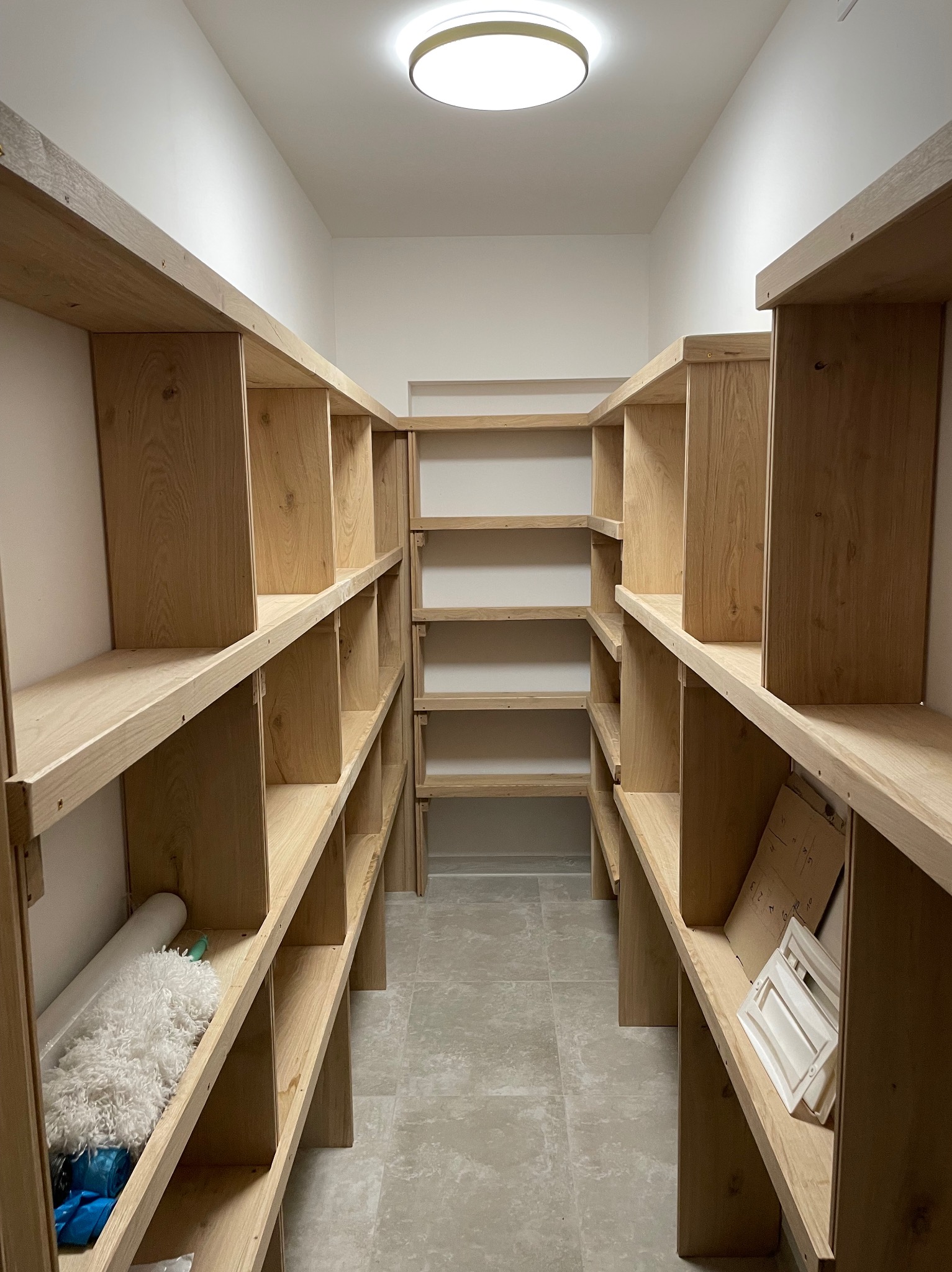 Wnętrze spiżarni z nowymi, jasnymi, drewnianymi półkami na całej ścianie, widok na korytarz, na jednej z półek leży mop, na innej kartony i białe ramki.