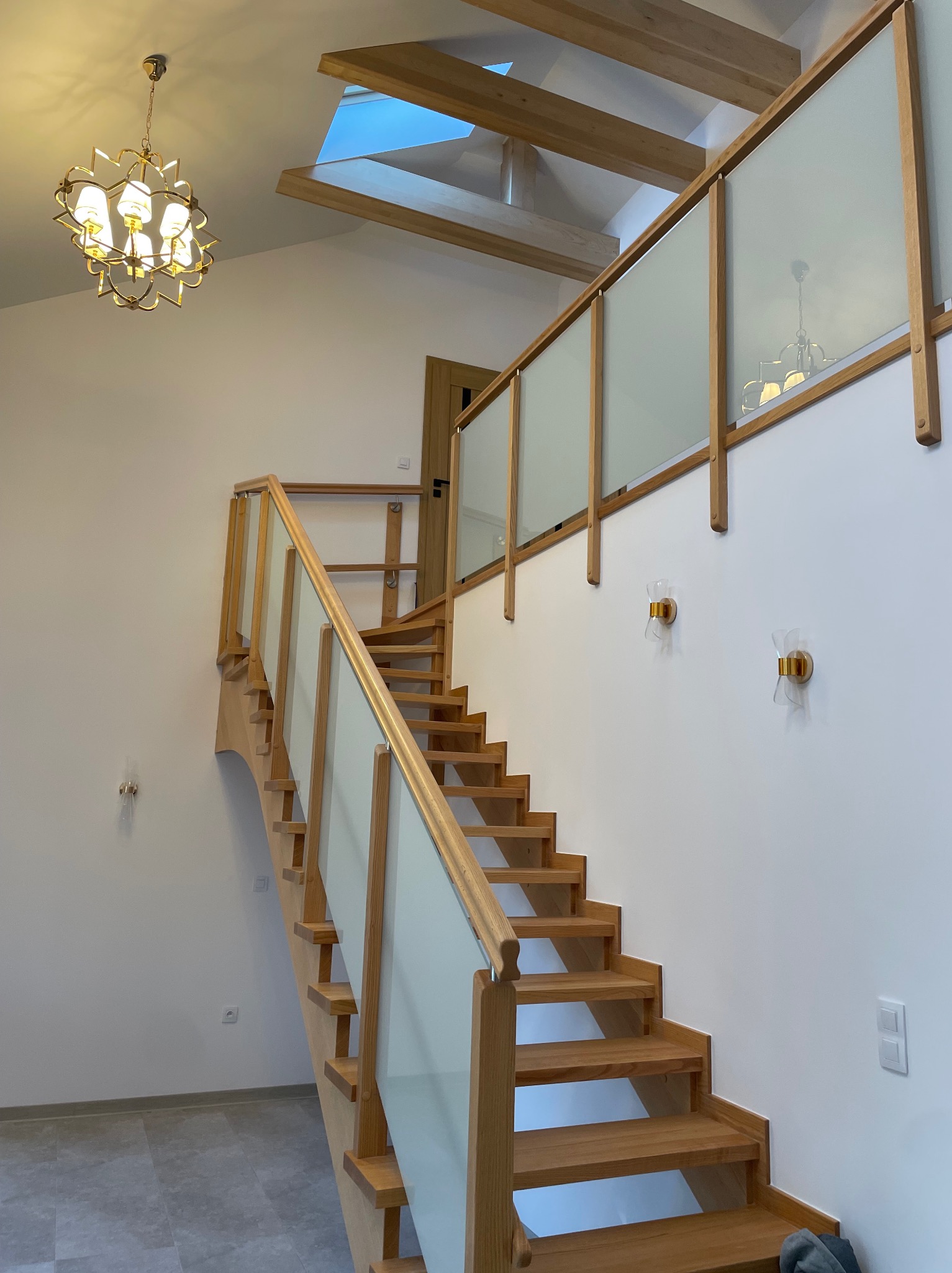 Jasne, drewniane schody z przeszkloną balustradą w nowoczesnym domu z oknem dachowym i ozdobnym żyrandolem.