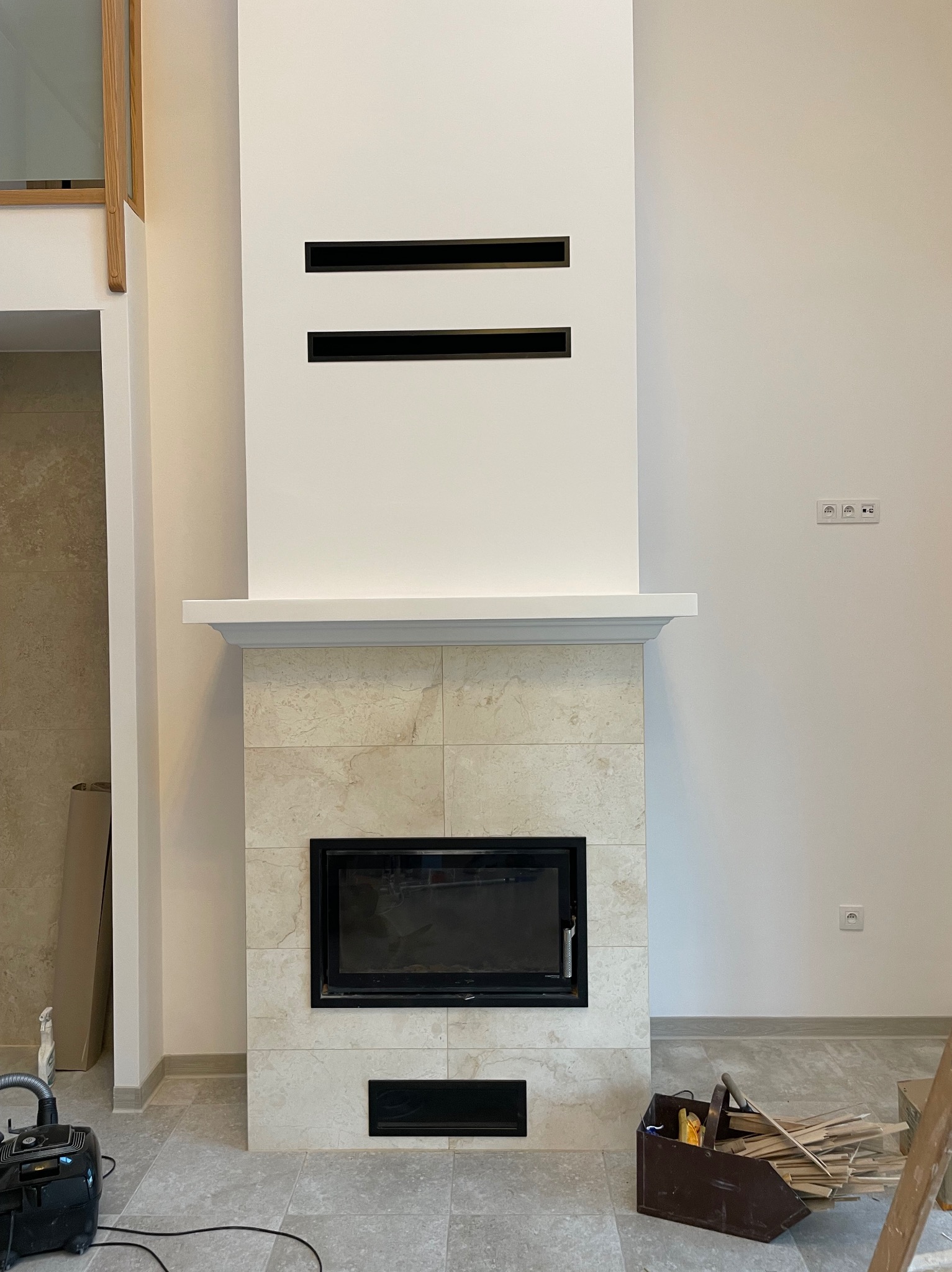 Nowoczesny kominek z obudową z jasnego kamienia, czarny wkład kominkowy, biała ściana z poziomymi szczelinami wentylacyjnymi, widoczny odkurzacz i skrzynka z narzędziami na podłodze.