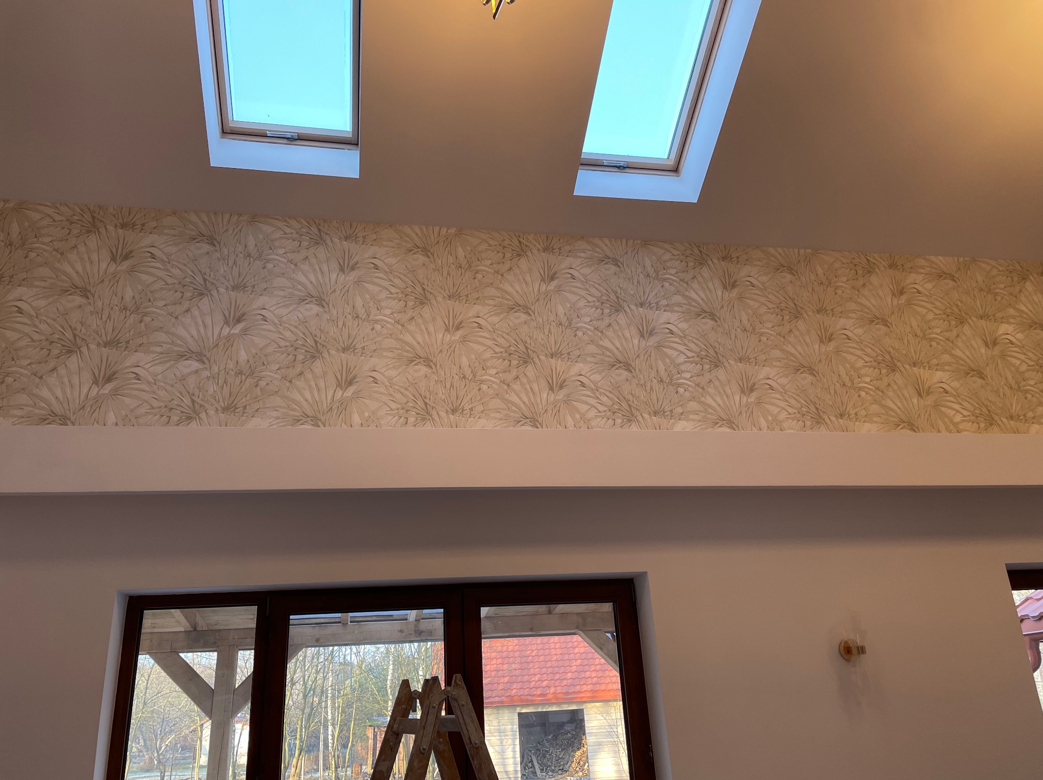 Ściana z wzorzystą tapetą w odcieniach beżu i brązu, widoczna nad oknem i schodami, oświetlona przez dwa okna dachowe, z widocznym fragmentem drabiny malarskiej.