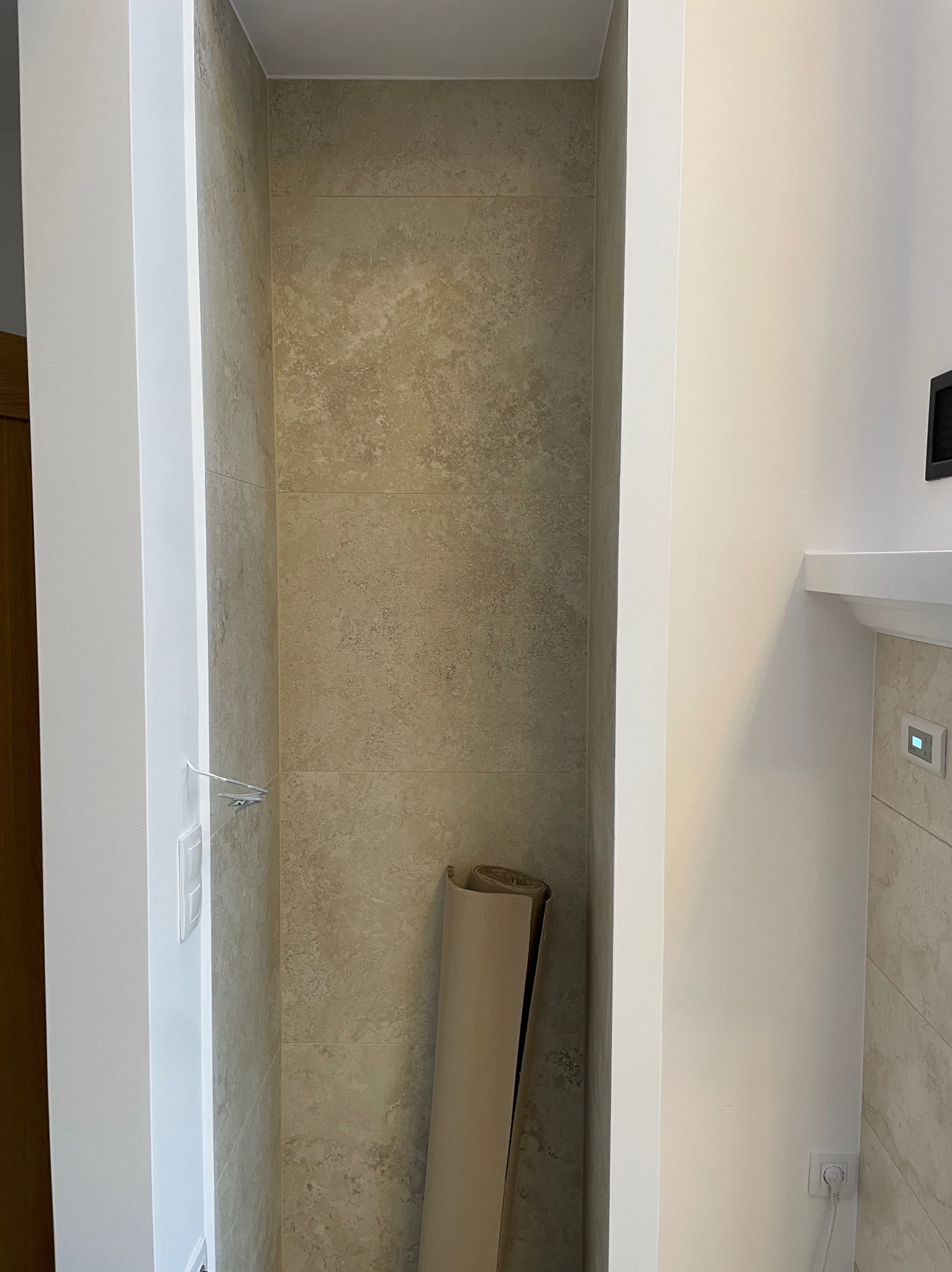 Wnęka w łazience wyłożona beżowymi płytkami imitującymi kamień, z rolką materiału zabezpieczającego podłogę lub wannę w rogu.