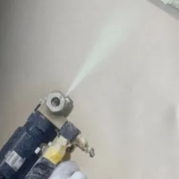 Natrysk pianki PUR na ścianę z bliska, widoczny pistolet aplikacyjny i dłoń w rękawicy. W tle ocieplona powierzchnia.