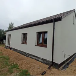 Nowo otynkowany, parterowy dom jednorodzinny z brązową dach&oacute;wką, brązowymi oknami i rynnami, widok z boku, teren wok&oacute;ł domu częściowo uprzątnięty, widoczna folia kubełkowa.