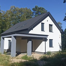 Arkadiusz Misiek - Nadzór i wsparcie budowlane - Nowoczesny, biały dom jednorodzinny z szarą stolarką okienną i dachem z blachy trapezowej, otoczony zielenią i drzewami.
