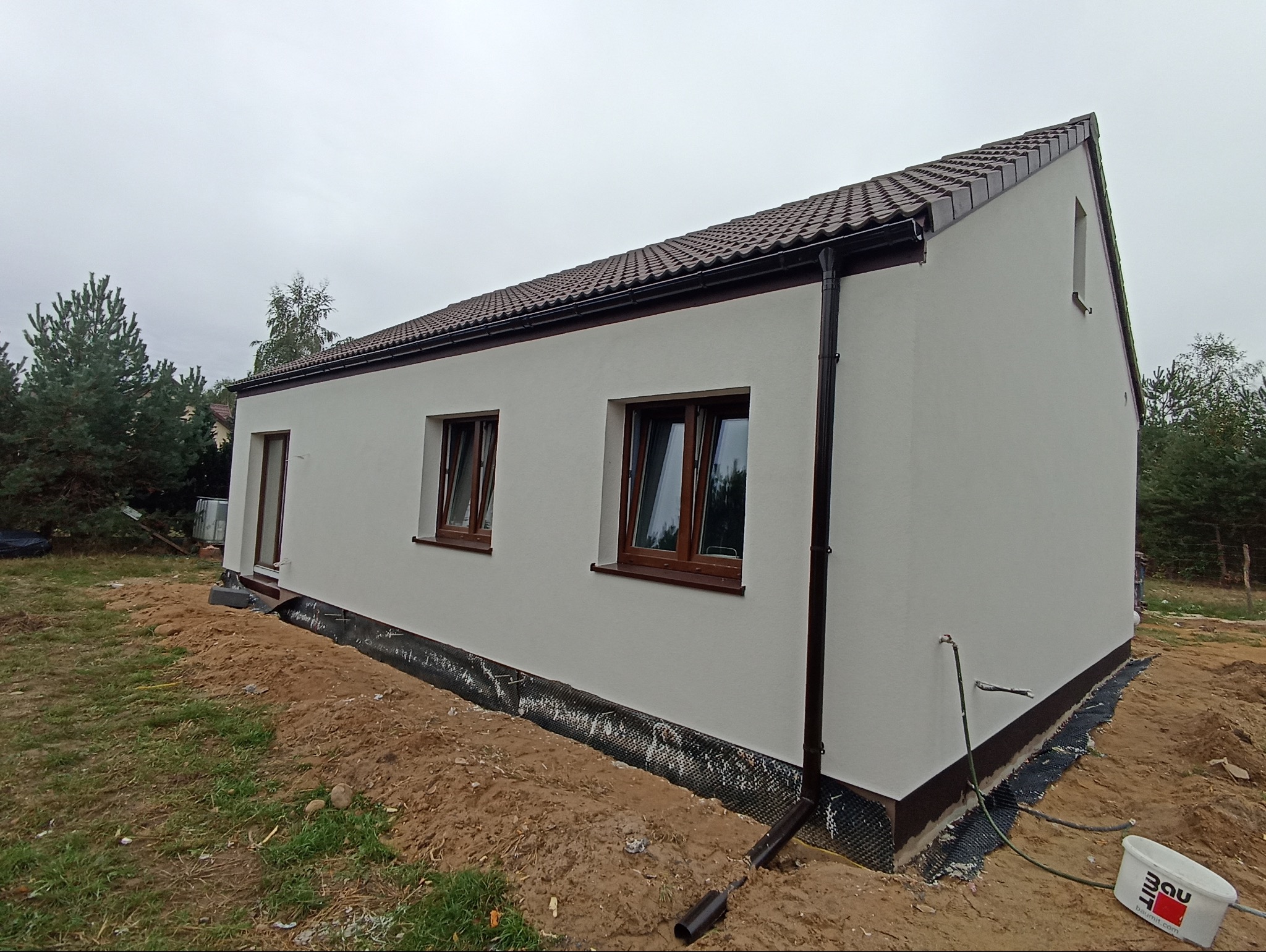 Nowo otynkowany, parterowy dom jednorodzinny z brązową dachówką, brązowymi oknami i rynnami, widok z boku, teren wokół domu częściowo uprzątnięty, widoczna folia kubełkowa.