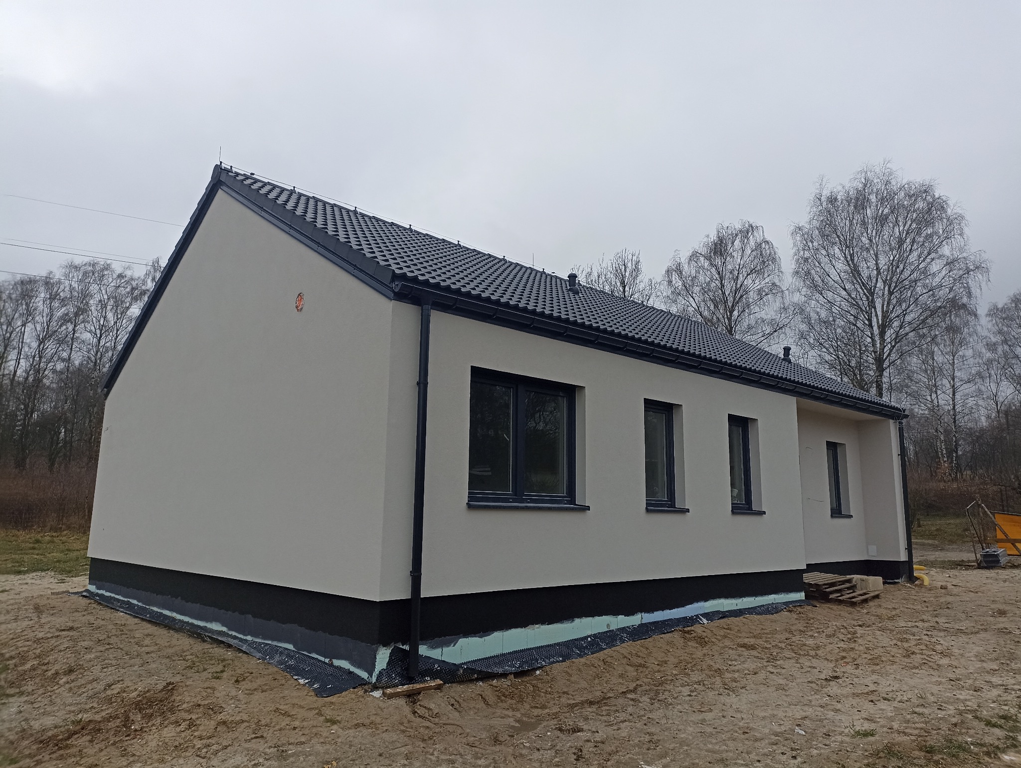 Nowo wybudowany, parterowy dom jednorodzinny z czarną dachówką i wykończoną elewacją w odcieniach szarości, widok z boku na tle drzew bez liści i pochmurnego nieba.