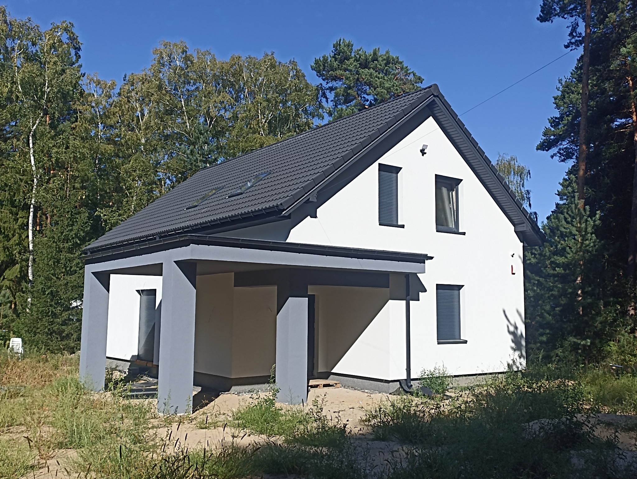 Nowoczesny, biały dom jednorodzinny z szarą stolarką okienną i dachem z blachy trapezowej, otoczony zielenią i drzewami.