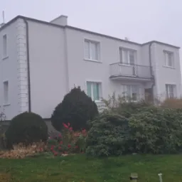 Dwupiętrowy dom z jasną elewacją, zdobieniami i balkonem, częściowo zasłonięty przez bujną roślinność ogrodową w pochmurny dzień.