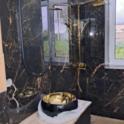 Nowoczesna łazienka w trakcie wykańczania: czarne płytki marmurowe z złotymi żyłami na ścianach, owalne lustro w czarnej ramie, złota armatura prysznicowa i czarno-złota umywalka nablatowa...