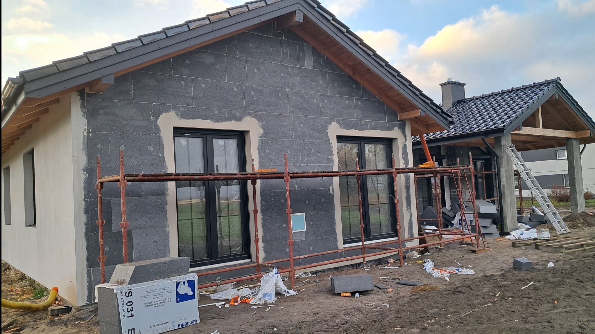 Dom w trakcie ocieplania elewacji: widoczne szare płyty izolacyjne, rusztowanie, okna z czarnymi ramami, częściowo otynkowana ściana i teren budowy z resztkami materiałów.
