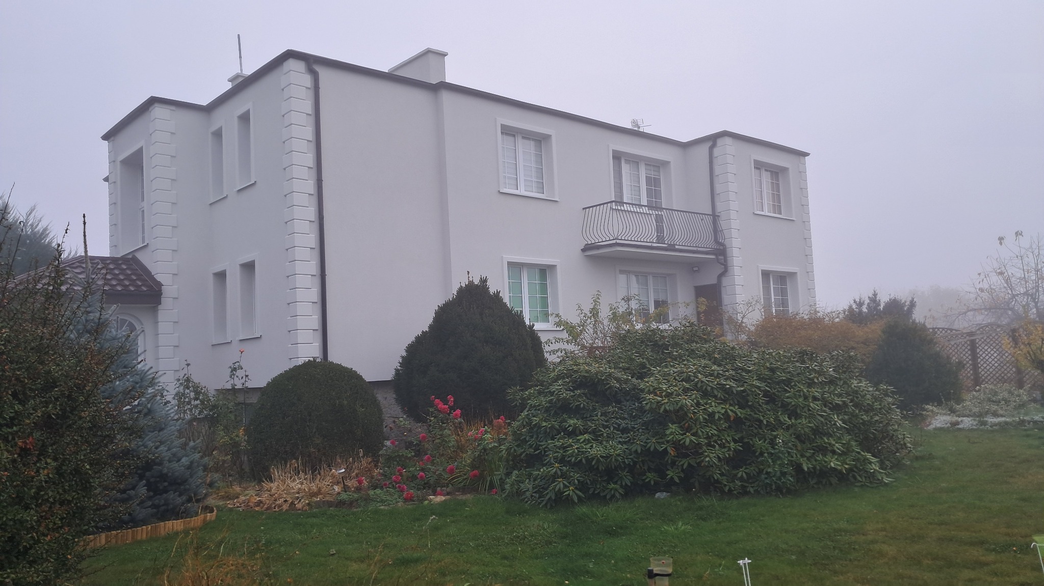 Dwupiętrowy dom z jasną elewacją, zdobieniami i balkonem, częściowo zasłonięty przez bujną roślinność ogrodową w pochmurny dzień.