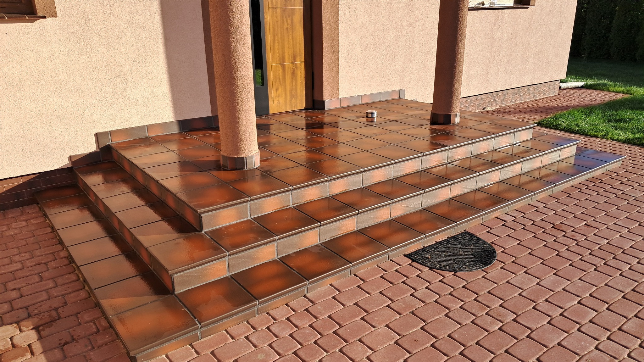 Błyszczące, brązowe płytki ceramiczne na schodach wejściowych do domu, z widocznymi kolumnami i wycieraczką przed drzwiami.