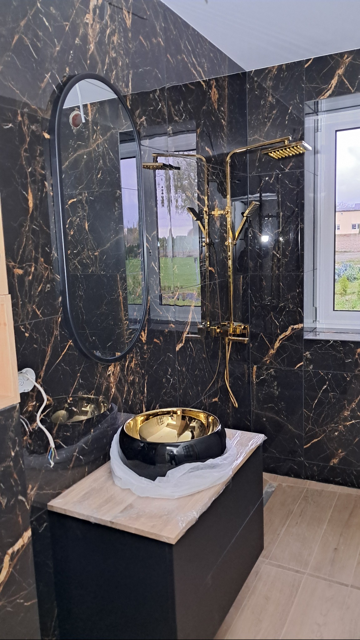 Nowoczesna łazienka w trakcie wykańczania: czarne płytki marmurowe z złotymi żyłami na ścianach, owalne lustro w czarnej ramie, złota armatura prysznicowa i czarno-złota umywalka nablatowa...