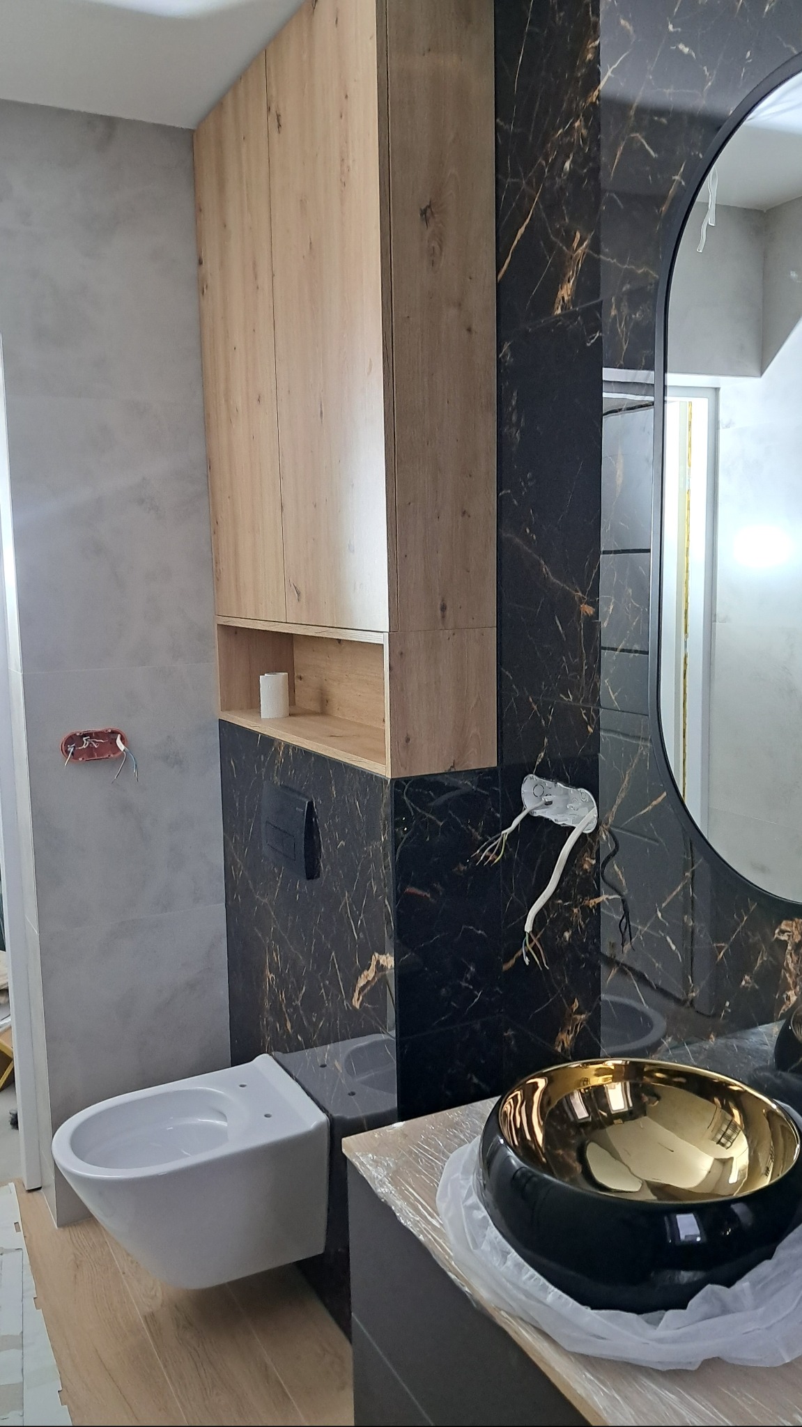 Nowoczesna łazienka w trakcie wykańczania: podwieszana toaleta, szafka z drewnianymi frontami, czarna marmurowa ściana z owalnym lustrem i złotą umywalką nablatową, widoczne przewody elektryczne.