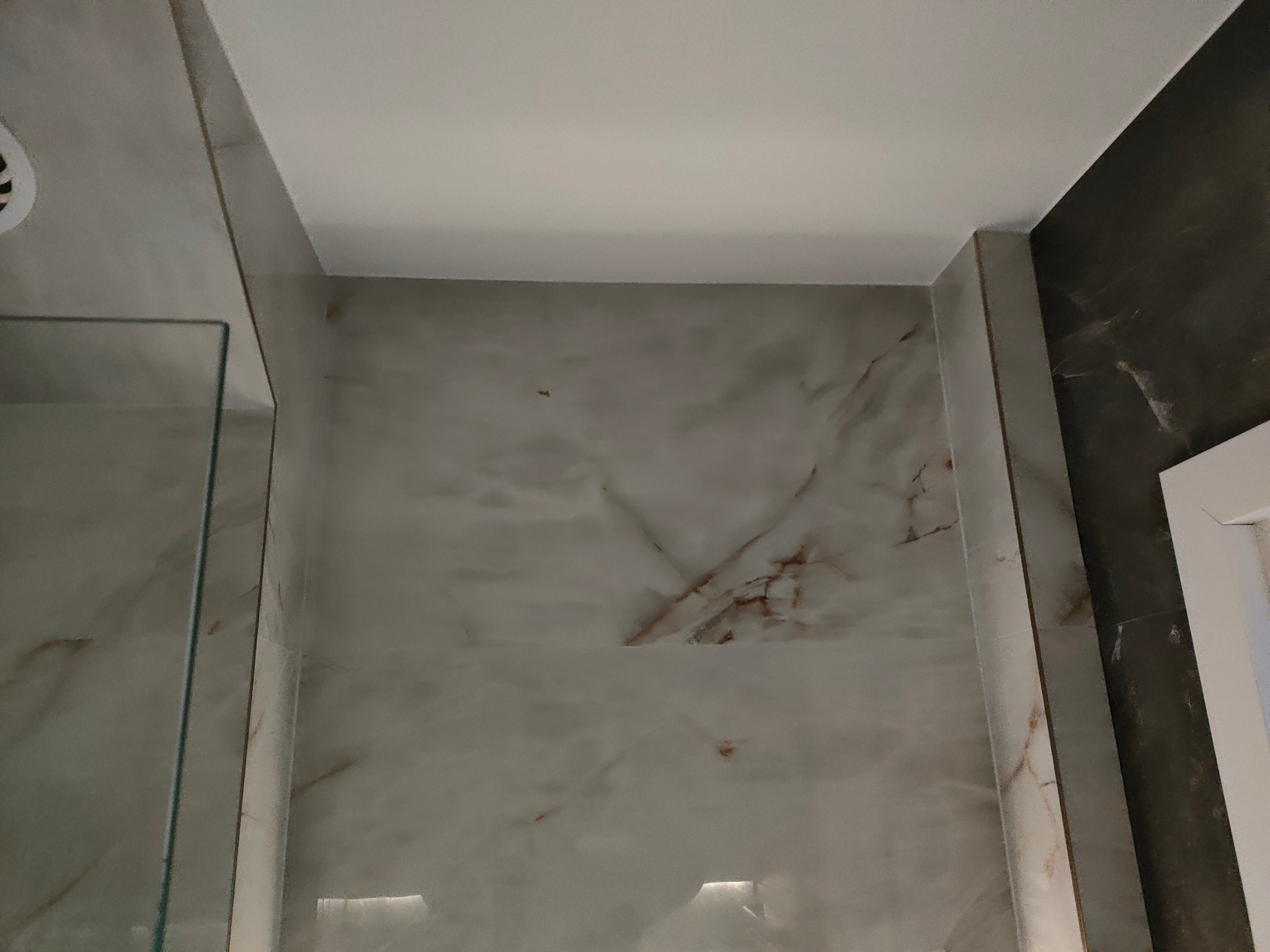 Wykończona ściana łazienki z marmurową okładziną, widoczna kabina prysznicowa z przezroczystego szkła i fragment sufitu. Minimalistyczny design z dbałością o detale.