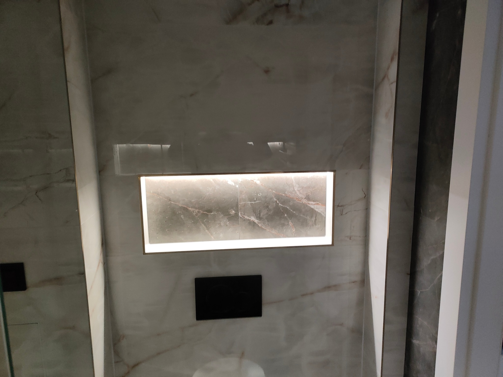 Narożna półka z podświetleniem LED w wykończonej marmurem łazience. Widoczny czarny przycisk spłuczki. Minimalistyczny design, ciepłe oświetlenie.