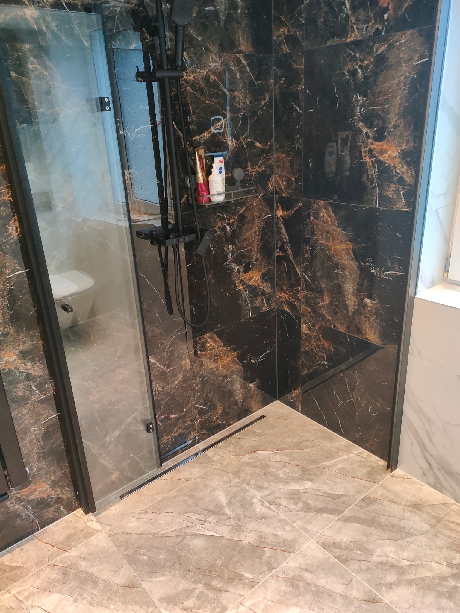 Nowoczesna kabina prysznicowa z czarną armaturą i marmurowymi płytkami w odcieniach brązu i beżu. W tle widoczna toaleta za szklaną ścianą.