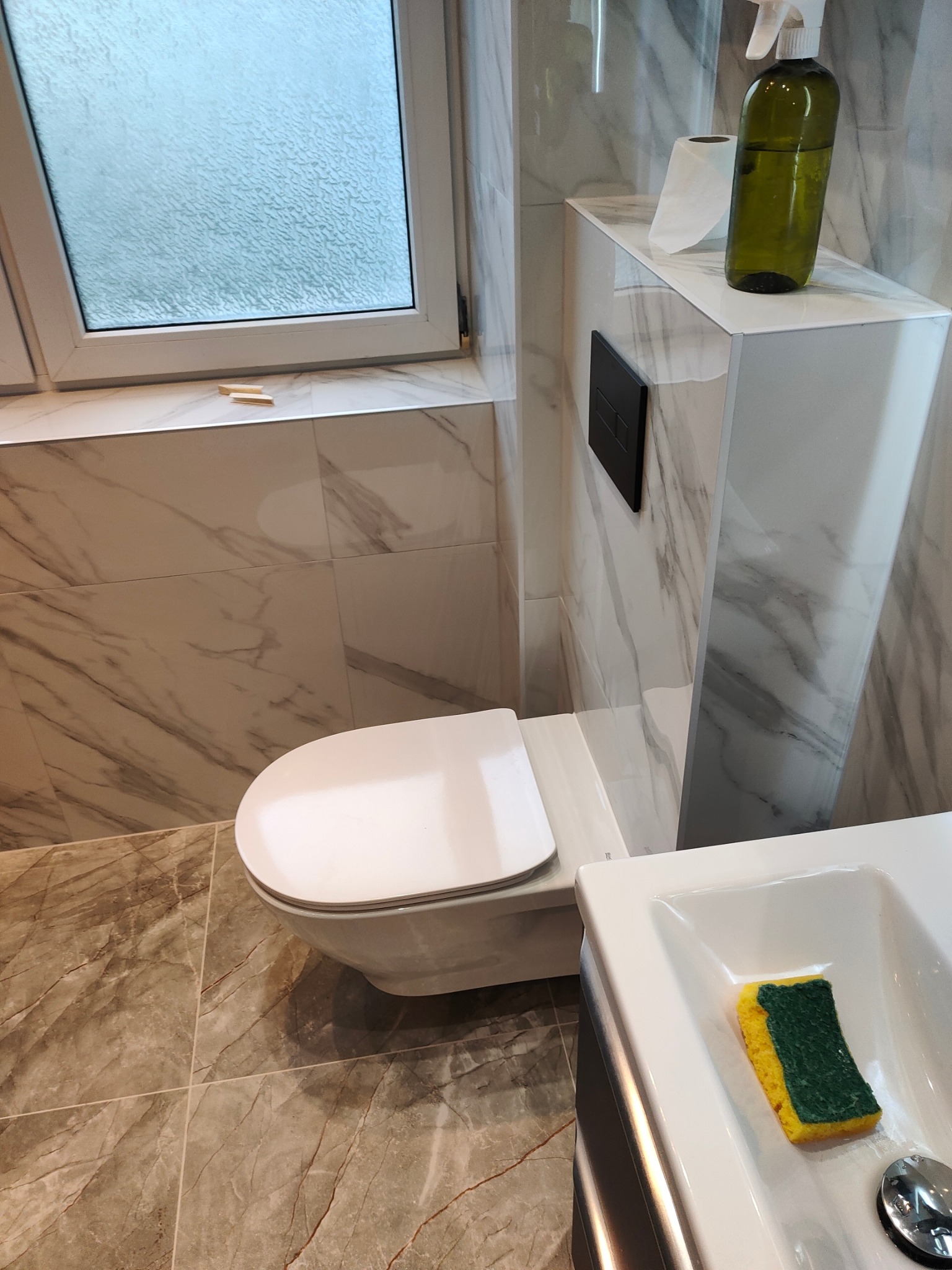 Nowoczesna łazienka z marmurowymi płytkami, podwieszaną toaletą i umywalką z gąbką. Okno z matową szybą zapewnia prywatność i doświetla wnętrze.