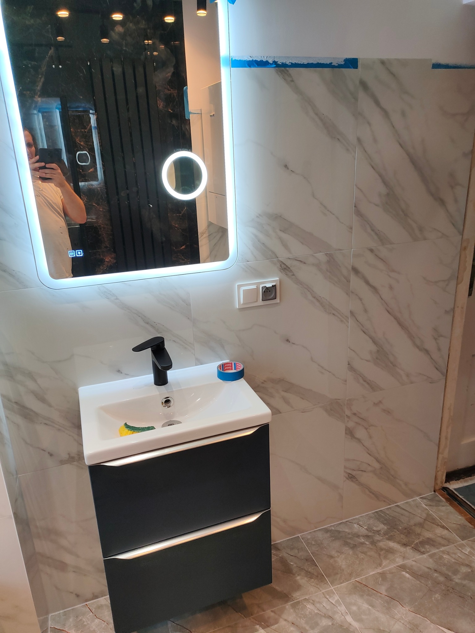 Nowoczesna łazienka z marmurowymi płytkami, umywalką z czarną szafką i lustrem z podświetleniem LED. Widoczna osoba robiąca zdjęcie w lustrze.