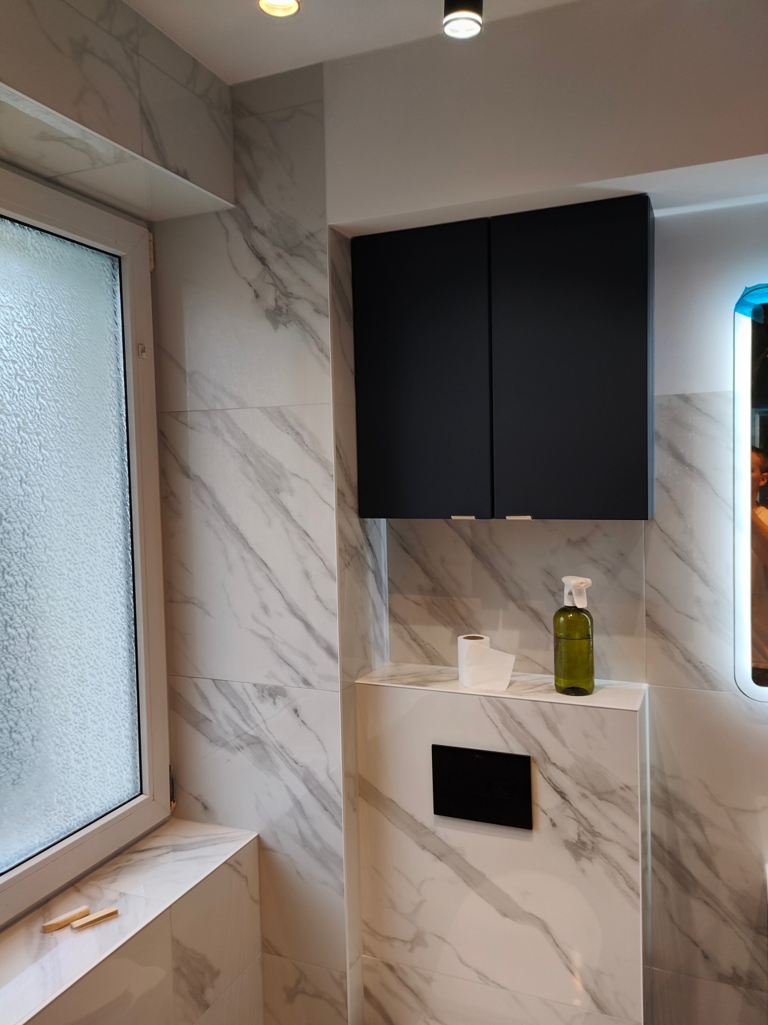 Wykończona łazienka z marmurowymi płytkami, czarną szafką nad toaletą i oknem z teksturowaną szybą. Na blacie papier toaletowy i butelka z dozownikiem.