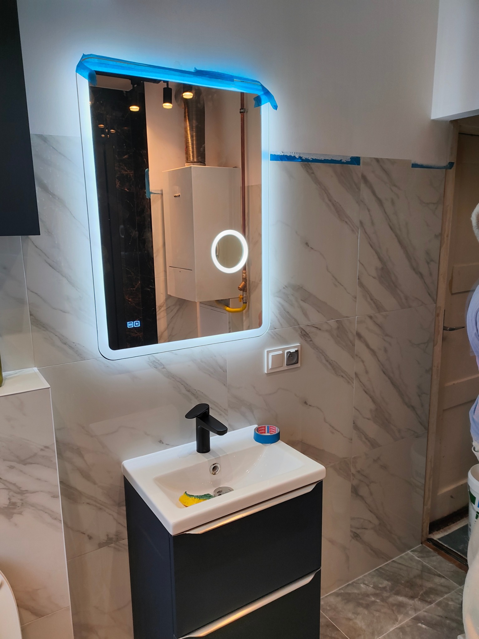 Ukończona łazienka z marmurowymi płytkami, szafką pod umywalką i lustrem z podświetleniem LED. Widoczny fragment kotła gazowego odbijający się w lustrze.