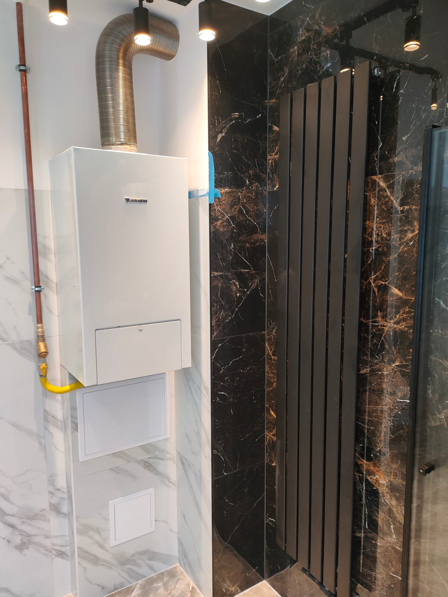 Nowoczesna łazienka z marmurowymi płytkami i grzejnikiem drabinkowym. Widoczny piec gazowy Junkers z instalacją. Minimalistyczny design, oświetlenie punktowe.