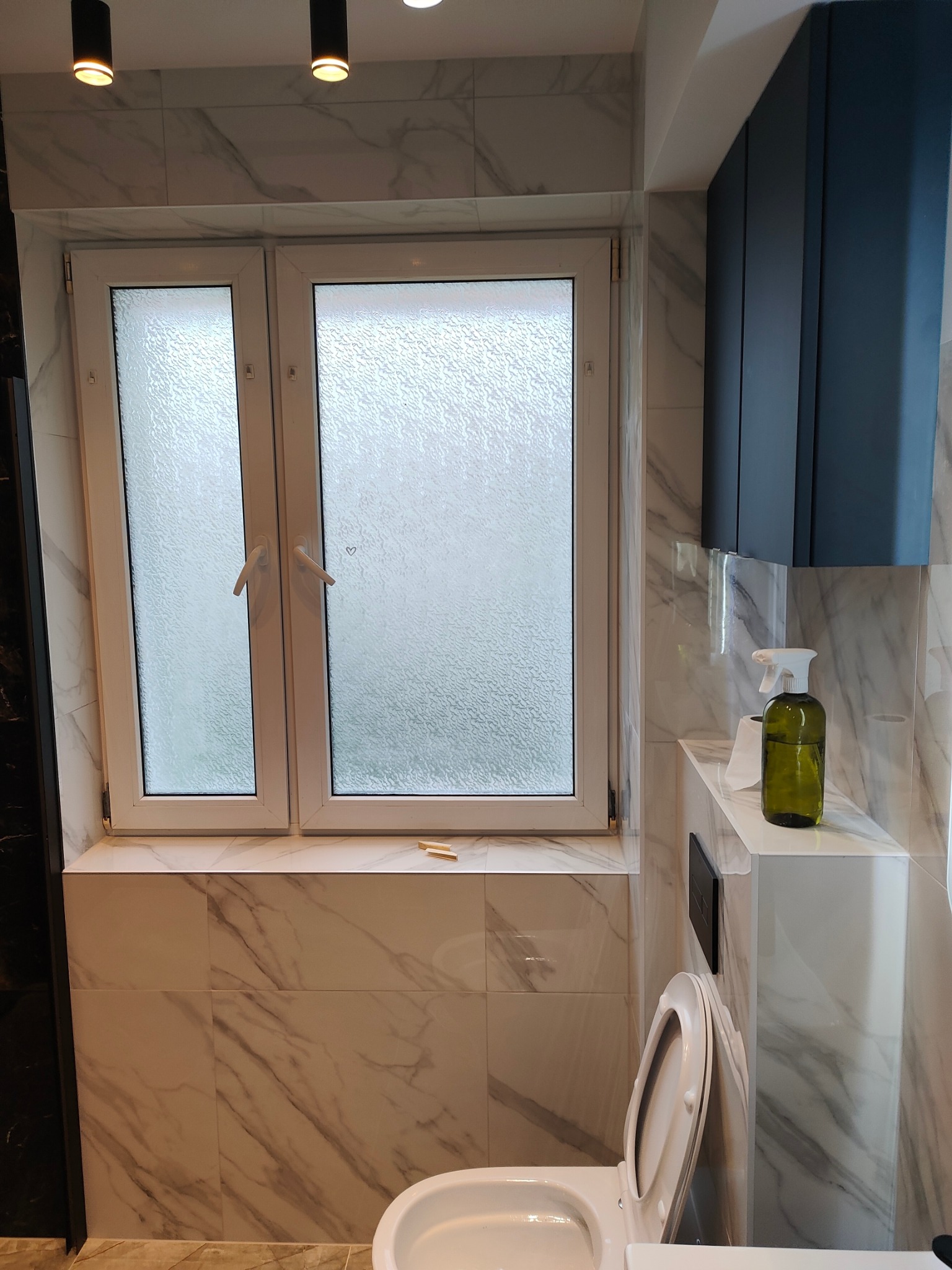 Wykończona łazienka z marmurowymi płytkami, zabudową szafek w kolorze granatowym i oknem z matową szybą. Widoczna toaleta i akcesoria łazienkowe.