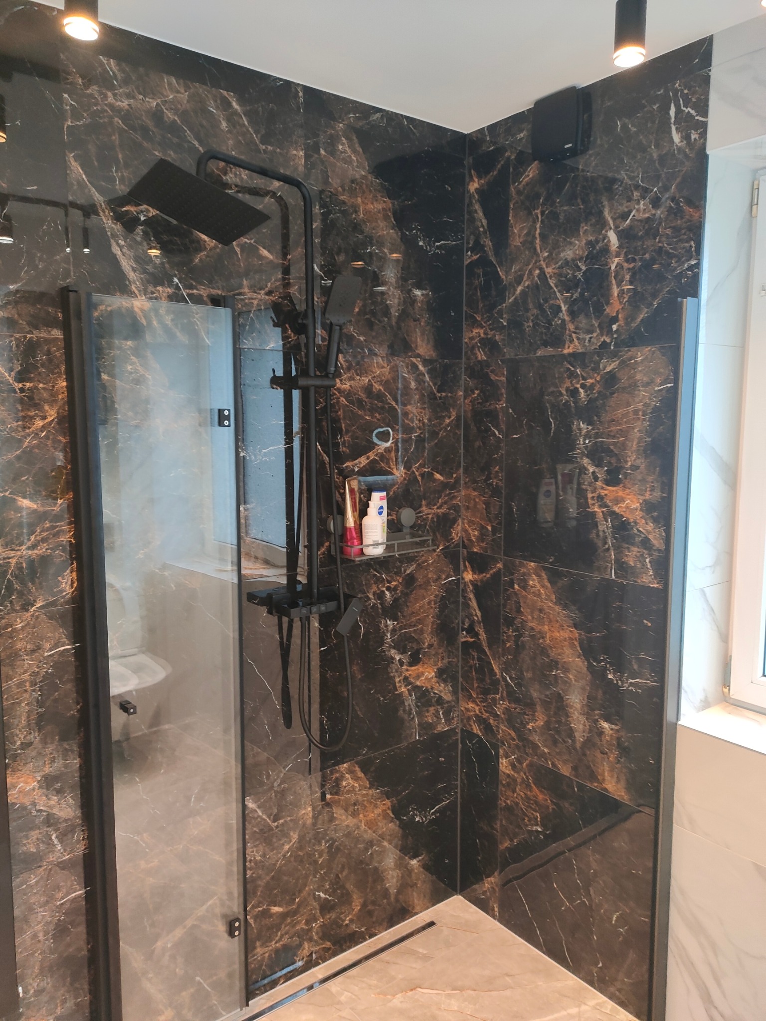 Nowoczesna kabina prysznicowa z czarną armaturą i płytkami imitującymi marmur w odcieniach brązu i czerni. Widoczne kosmetyki na półce, szklane drzwi kabiny i oświetlenie punktowe.