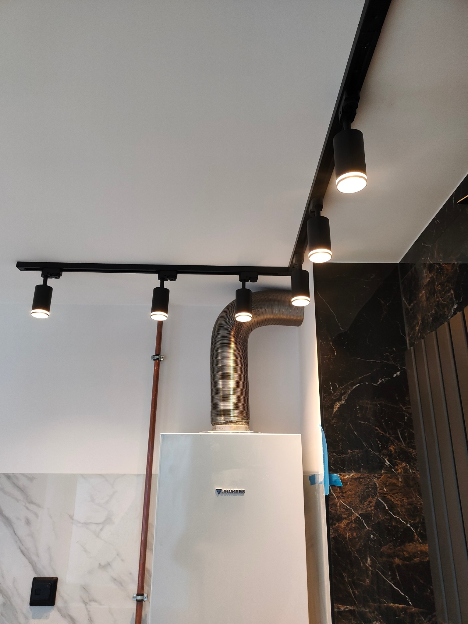 Biały piec gazowy Junkers z rurą spiro i miedzianą instalacją, oświetlony czarnymi reflektorami szynowymi na tle marmurowych płytek w łazience.