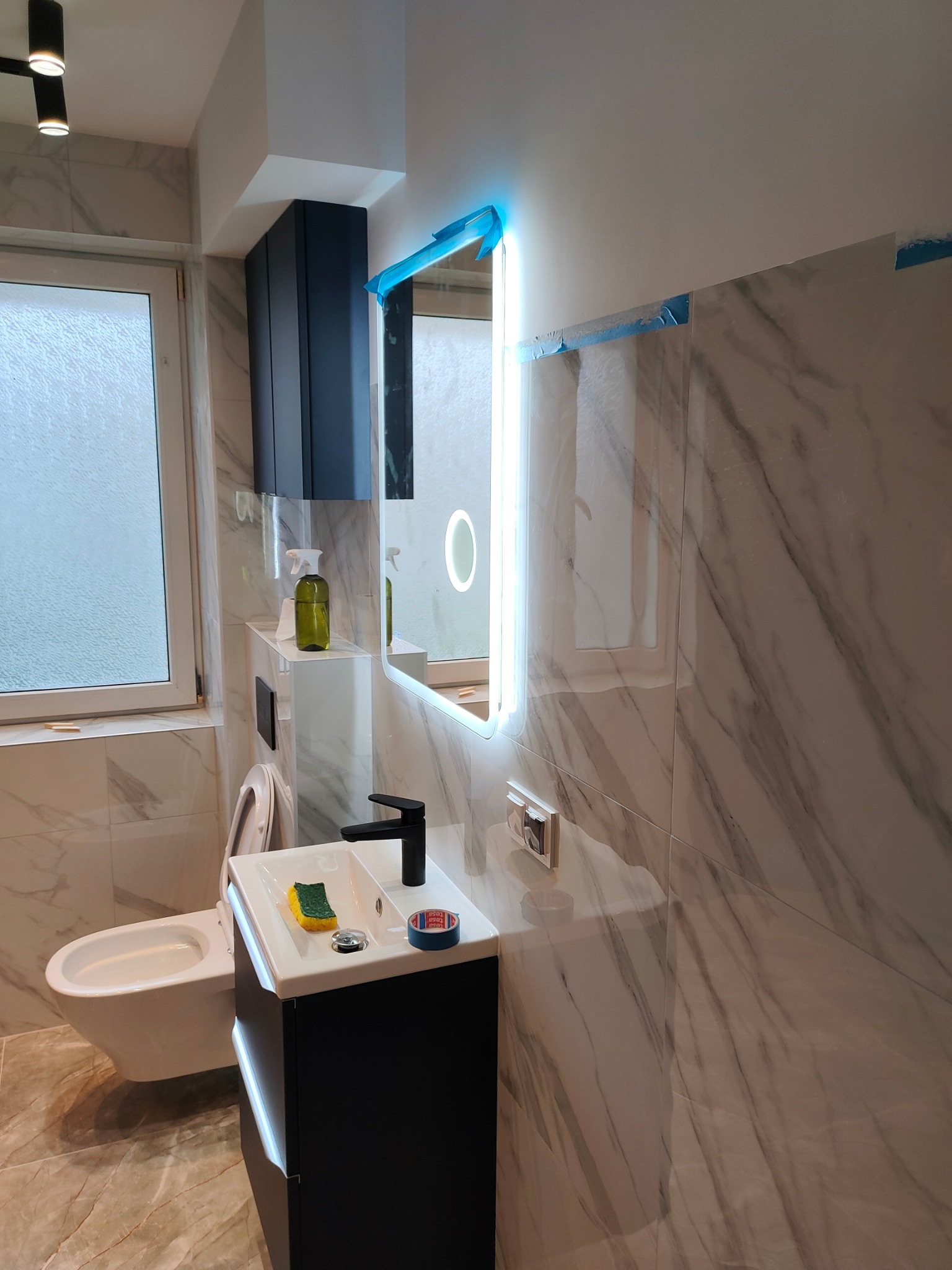 Nowoczesna łazienka z marmurowymi płytkami, podświetlanym lustrem i czarną armaturą. Fragmenty zabezpieczeń folią wskazują na świeżo zakończony remont.