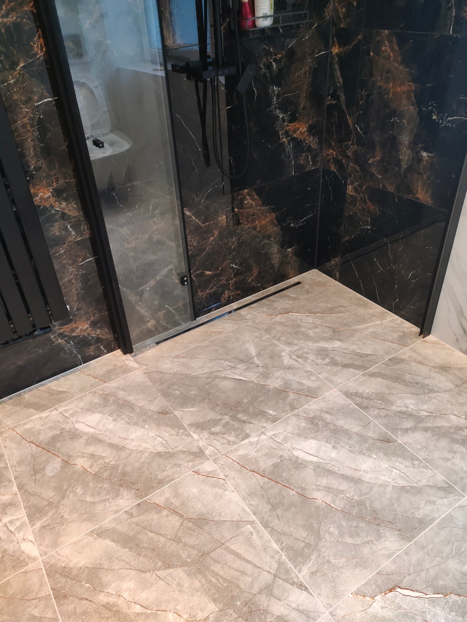 Nowoczesna łazienka z czarnymi płytkami imitującymi marmur na ścianach i jasnymi płytkami podłogowymi. Widoczna kabina prysznicowa z czarnymi elementami.