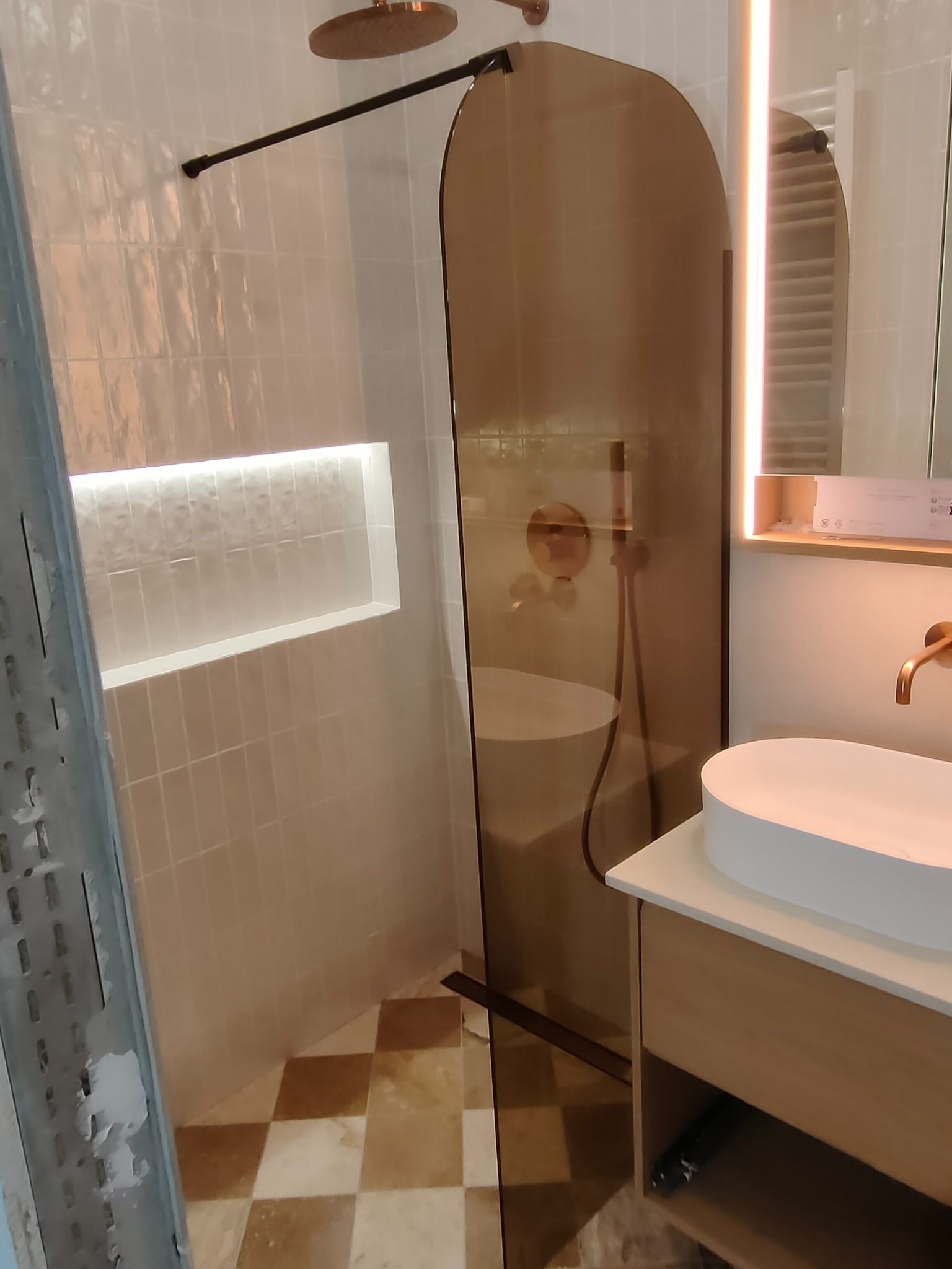 Nowoczesna łazienka z kabiną prysznicową typu walk-in, beżowymi płytkami i podświetlaną wnęką. Umywalka nablatowa na szafce z drewnopodobnym wykończeniem.