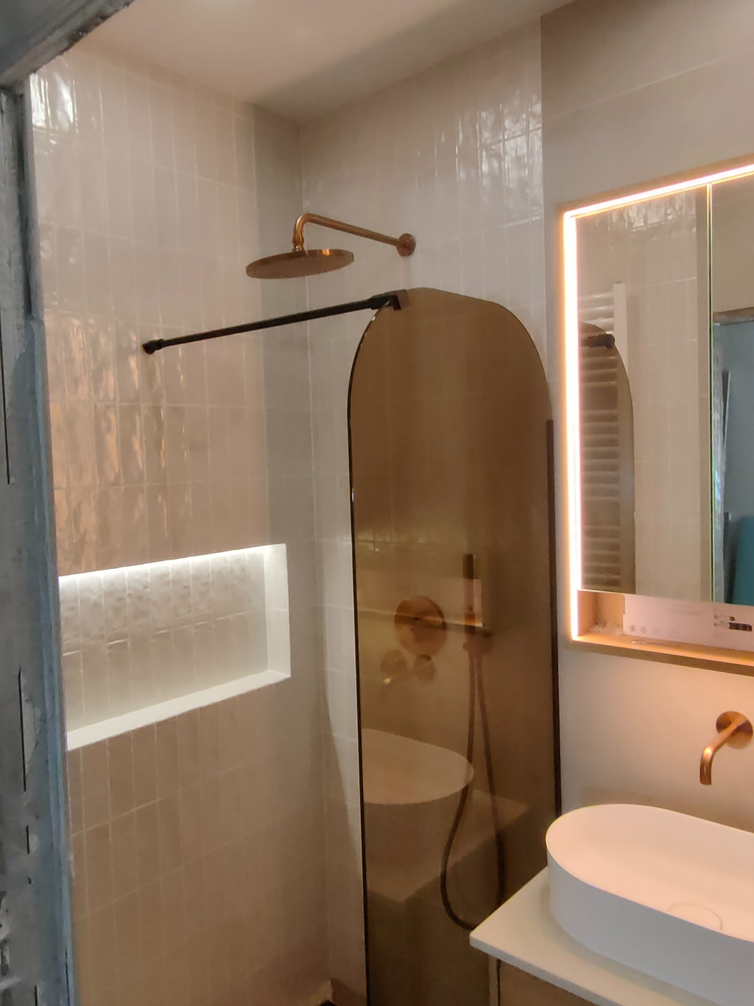 Nowoczesna łazienka z beżowymi płytkami, złotą armaturą i kabiną prysznicową z brązowego szkła. W ścianie wnęka z oświetleniem LED. Lustro z podświetleniem.