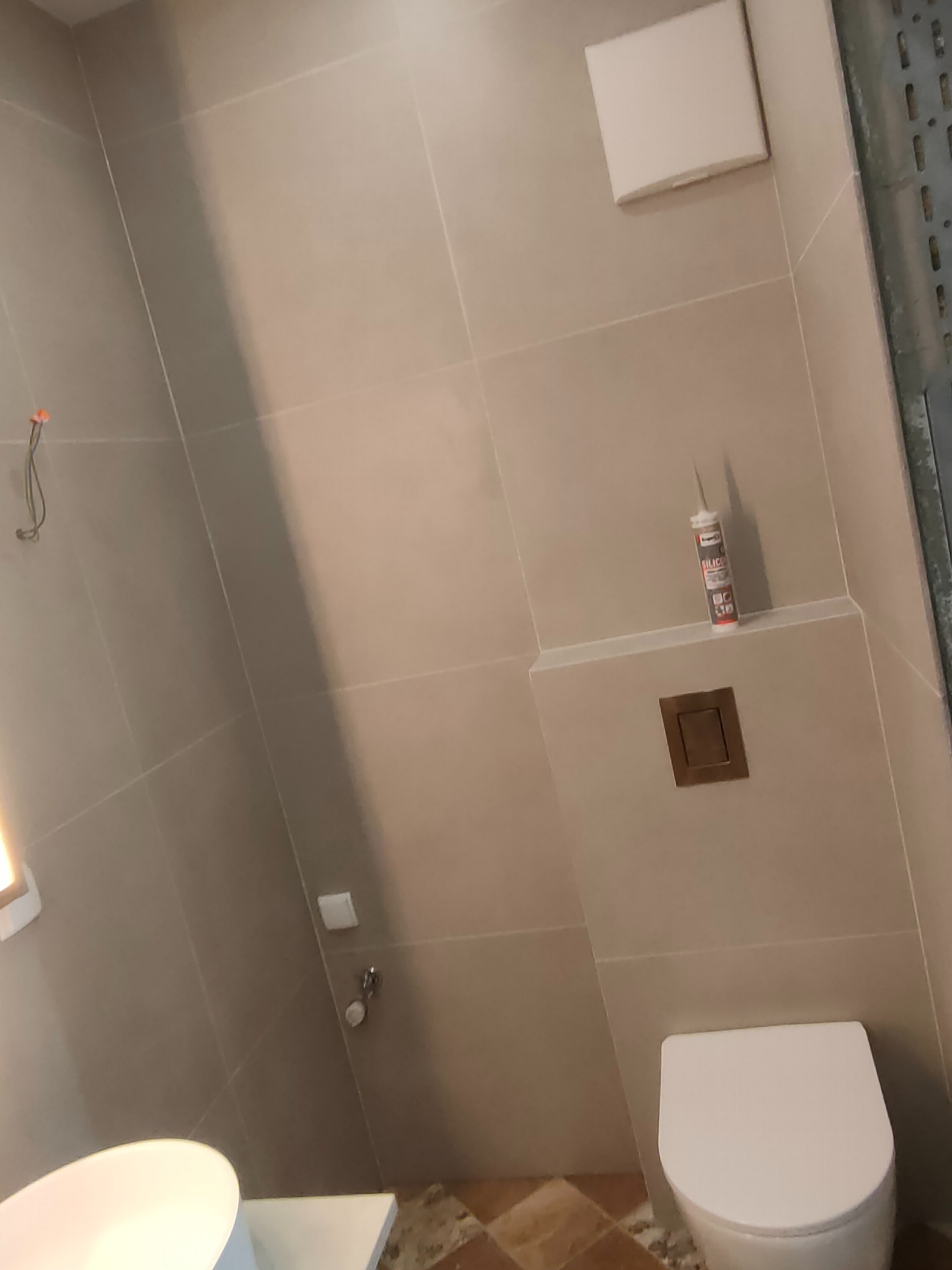 Beżowa łazienka w trakcie wykańczania: biała toaleta, umywalka, płytki, wentylator, silikon. Widoczne narzędzia i elementy instalacji. Ukończony etap prac.