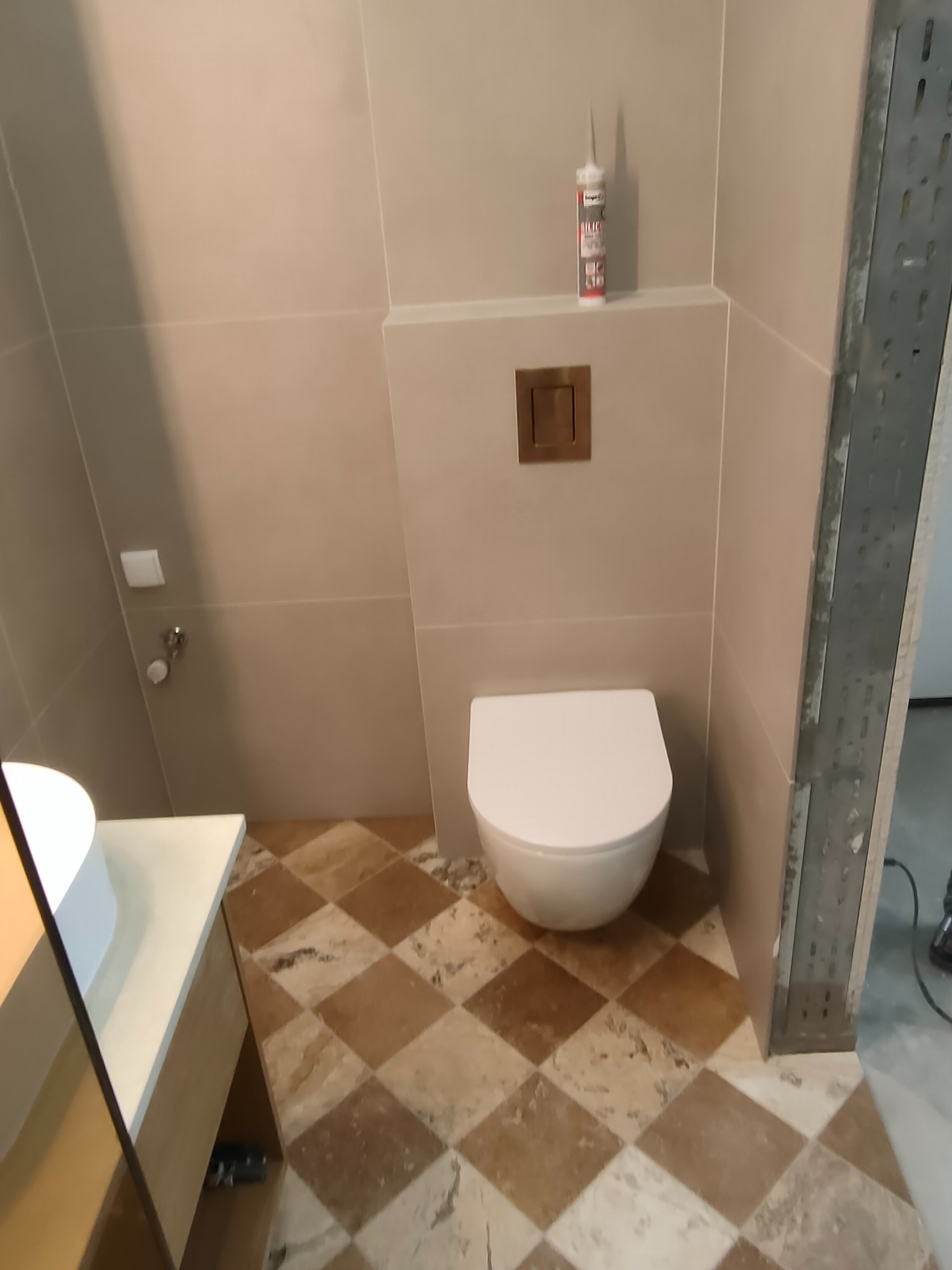 Wykończona łazienka z beżowymi płytkami, toaletą i umywalką. Na podłodze płytki w brązowo-beżową szachownicę. Widoczna tuba silikonu na półce.