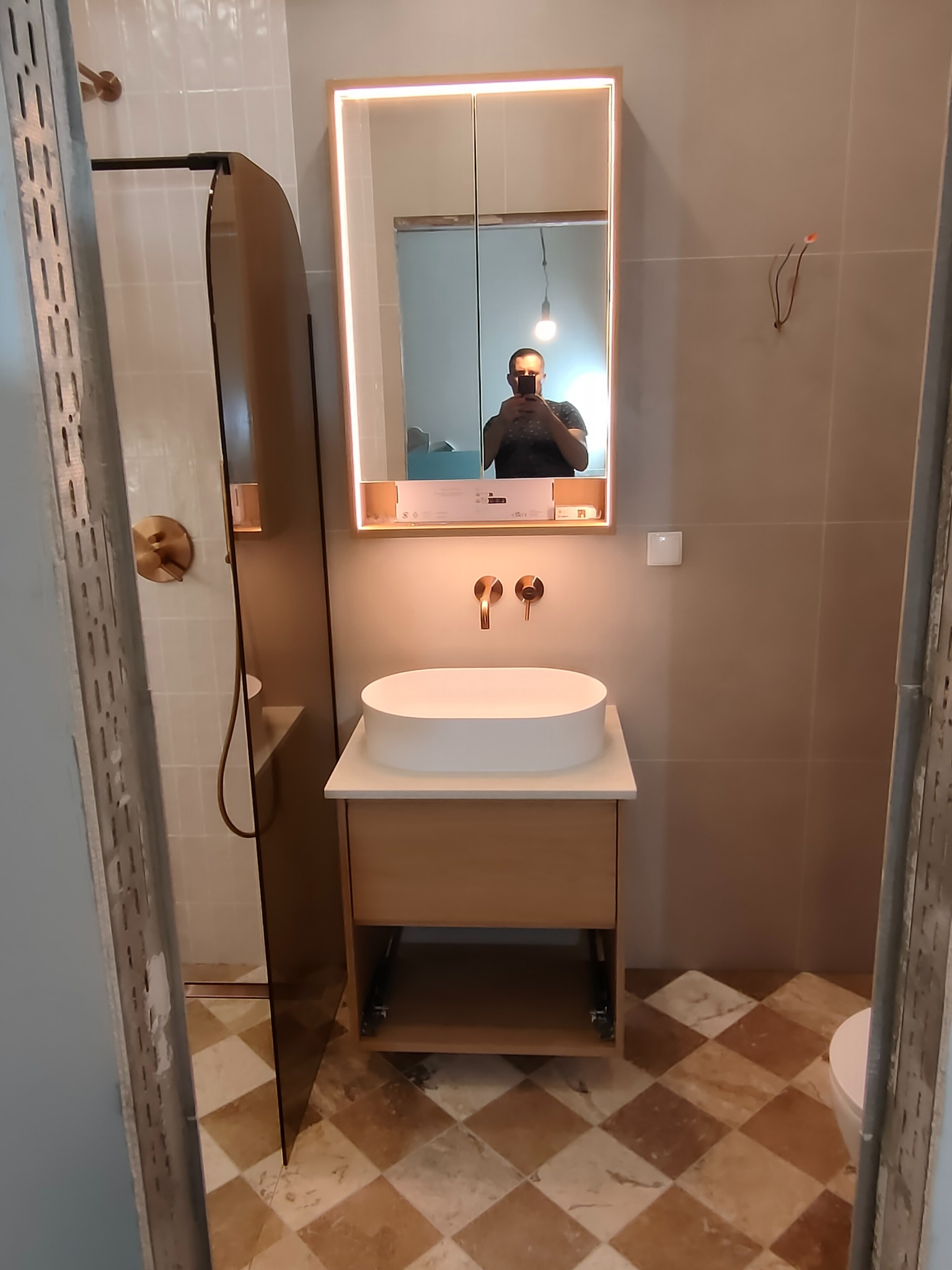 Nowoczesna łazienka z owalną umywalką na szafce z jasnego drewna, podświetlanym lustrem i kabiną prysznicową z brązowymi elementami. Na podłodze płytki w romby.