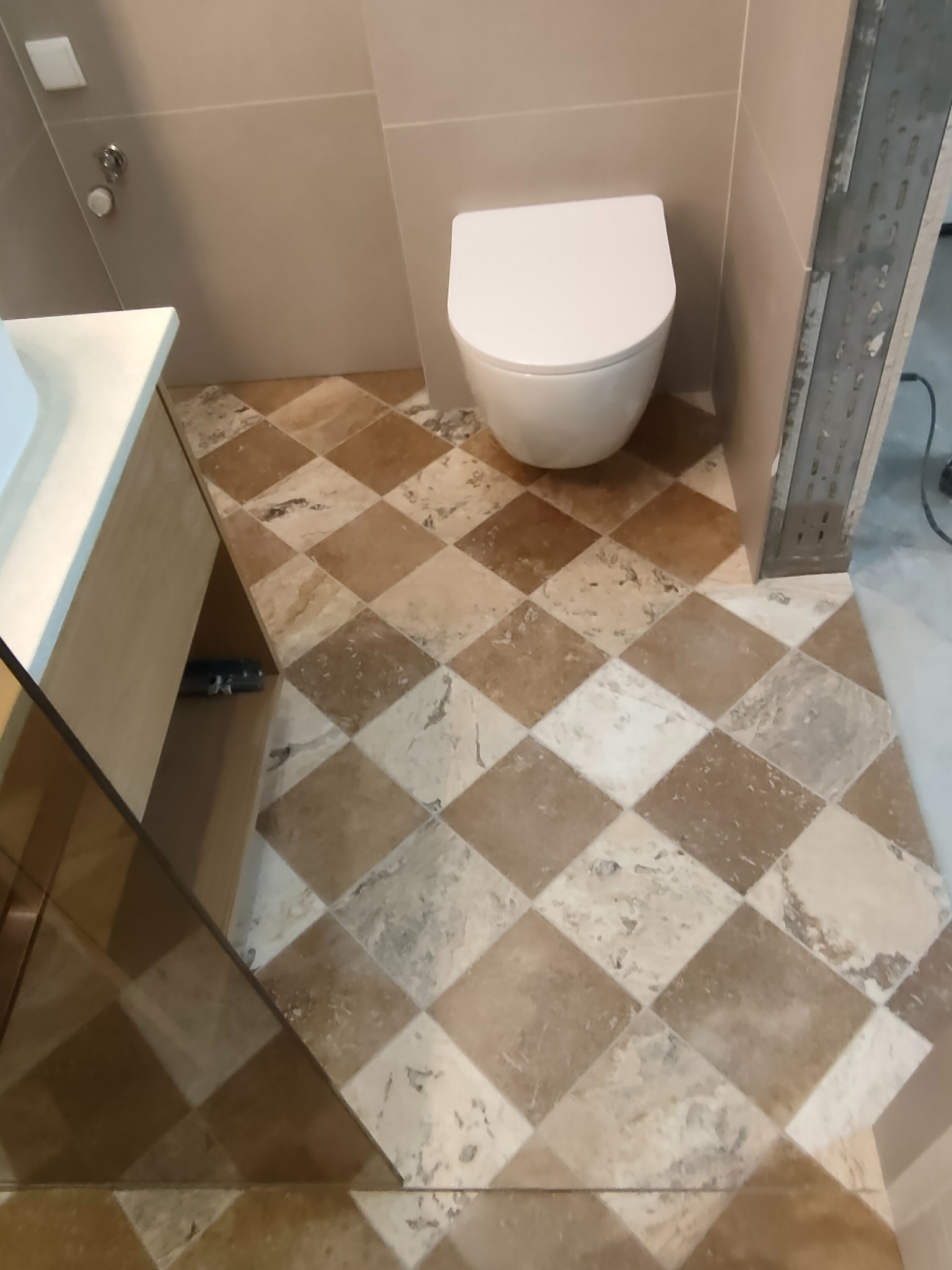 Ukończona łazienka z wzorzystą, romboidalną podłogą z płytek w odcieniach beżu i brązu, białą toaletą i częściowo widoczną szafką pod umywalkę.