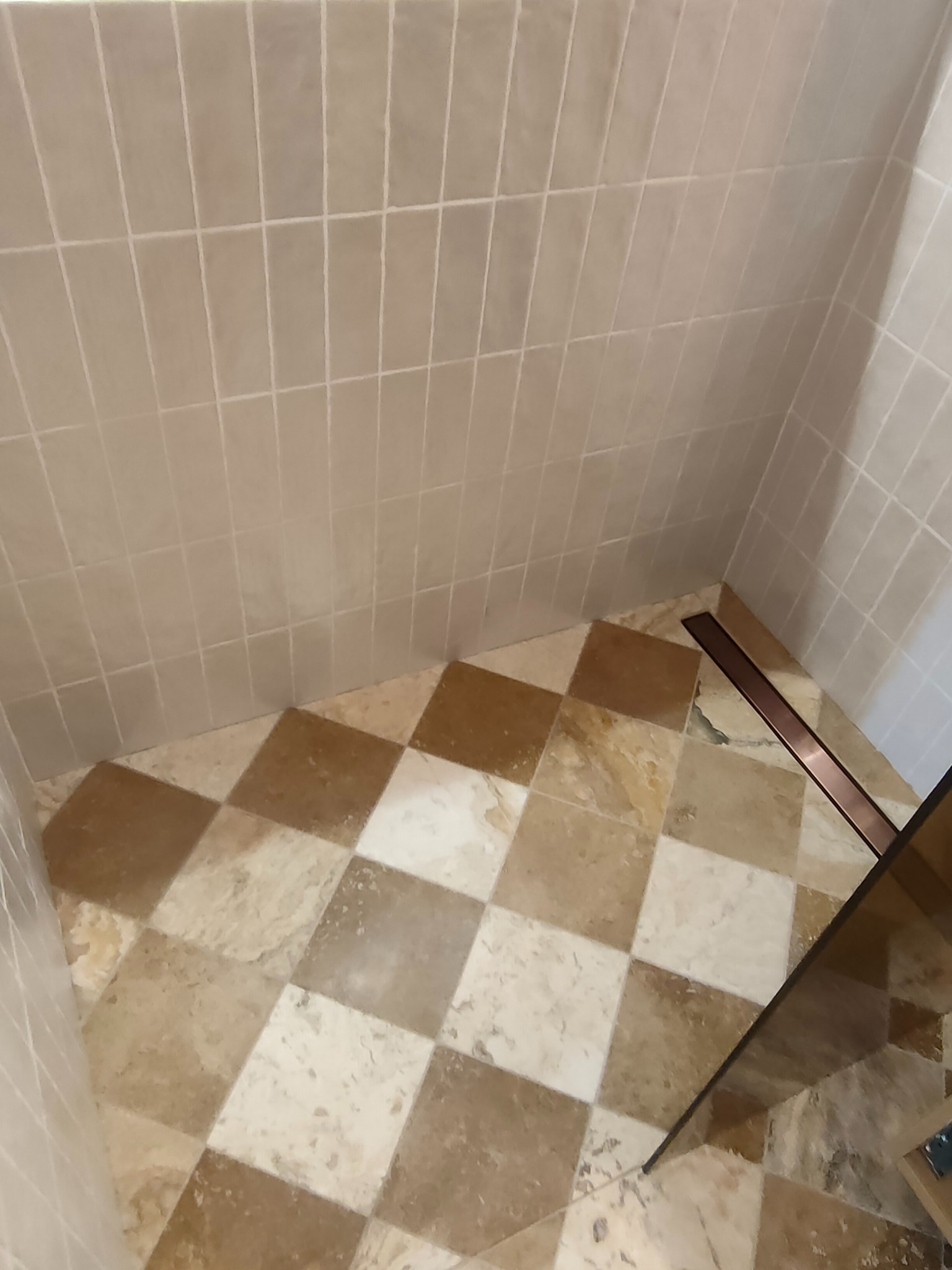 Beżowa łazienka z kwadratową podłogą w szachownicę i odpływem liniowym. Ściany wyłożone pionowymi, prostokątnymi płytkami w odcieniach beżu. Widok z góry na fragment kabiny.