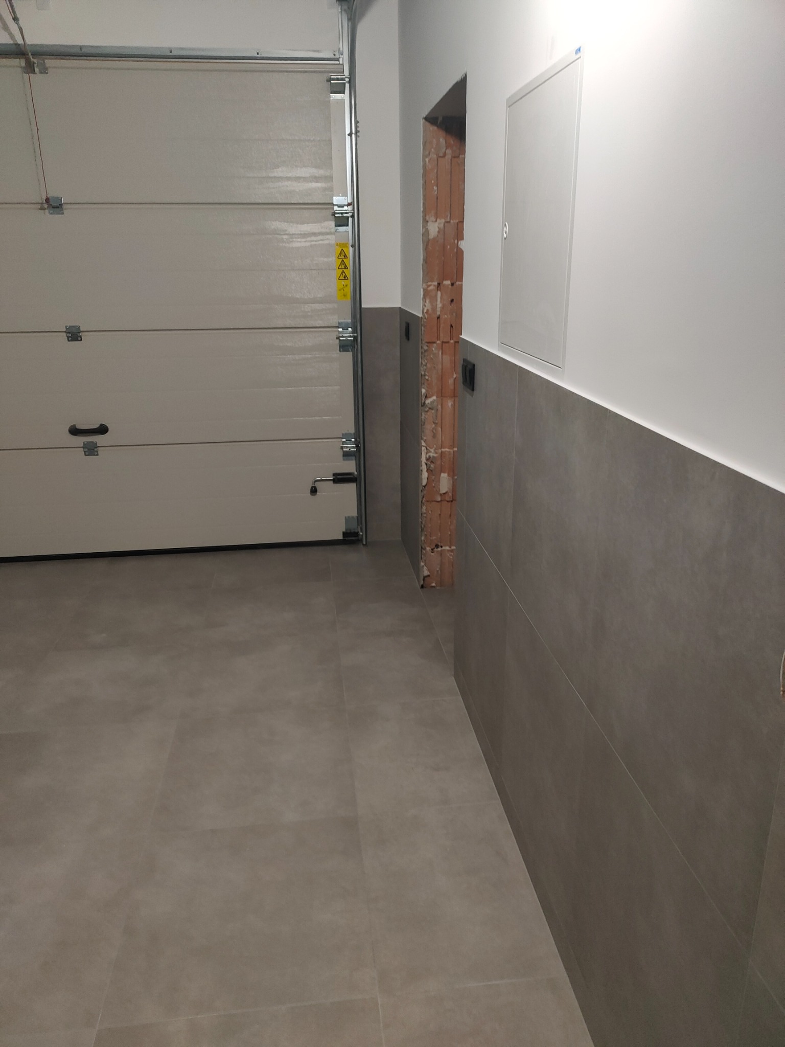 Wykończone płytki ścienne i podłogowe w korytarzu z otwartą bramą garażową. Widoczny fragment ściany z cegły w miejscu planowanego przejścia.
