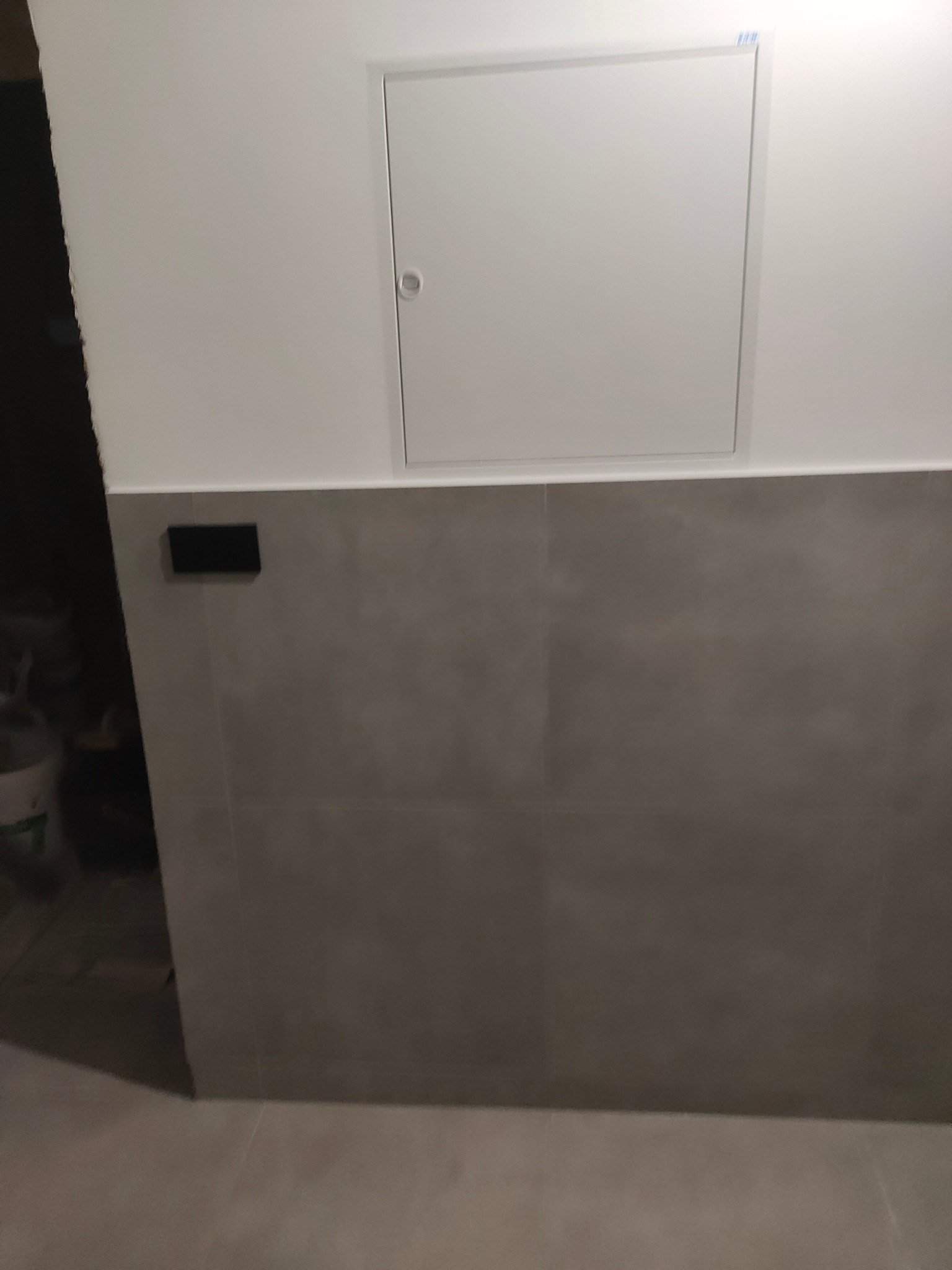 Szare płytki ścienne do połowy wysokości z białymi ścianami i drzwiczkami rewizyjnymi. Widoczny fragment podłogi i czarny włącznik.
